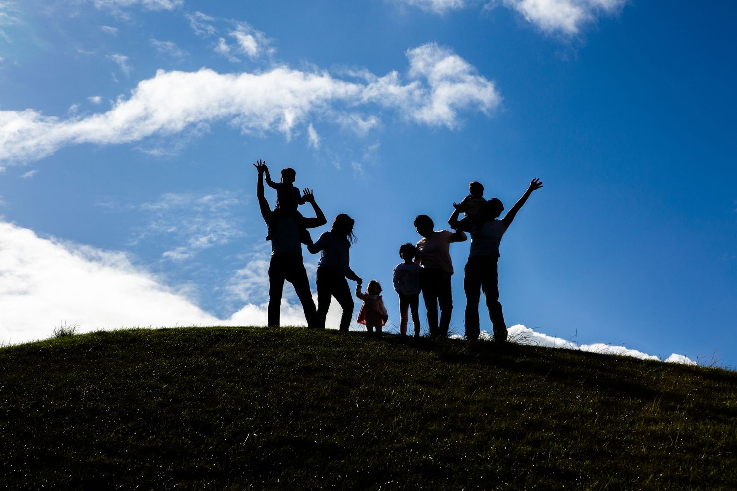 Silhouette of a joyful family with children standing on a grassy hill against a bright blue sky.