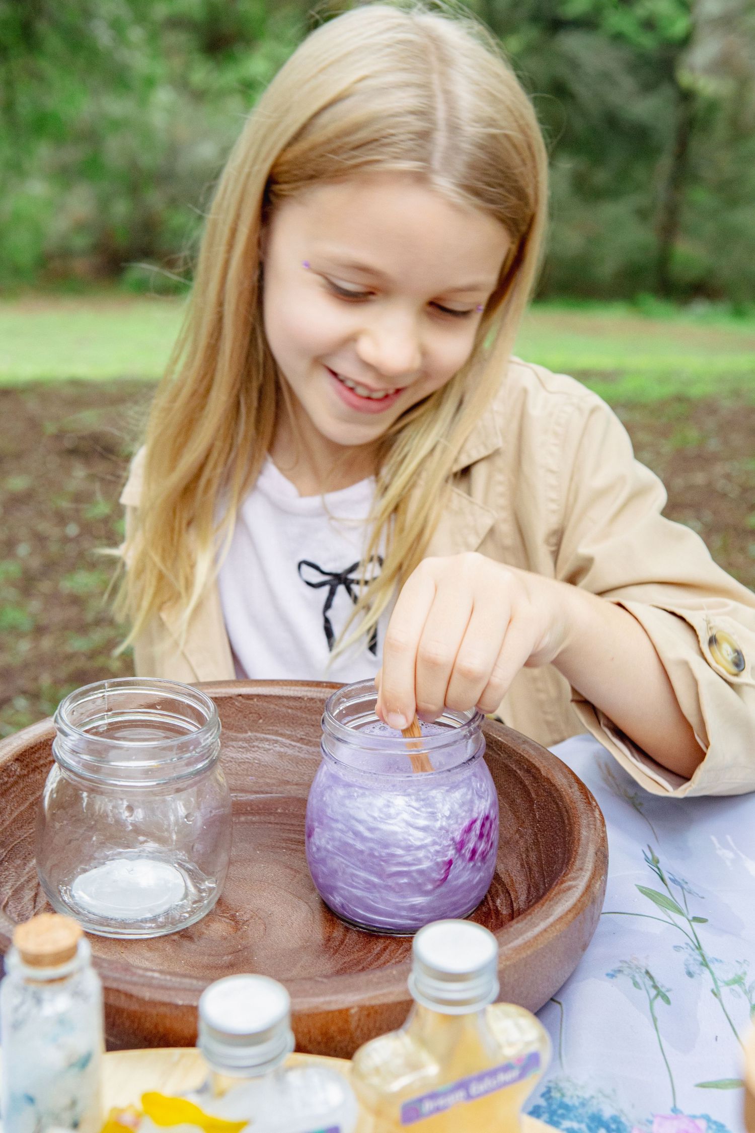 Smiling blonde girl dipping finger into purple liquid in jar on wooden tray outdoors, science experiment activity.