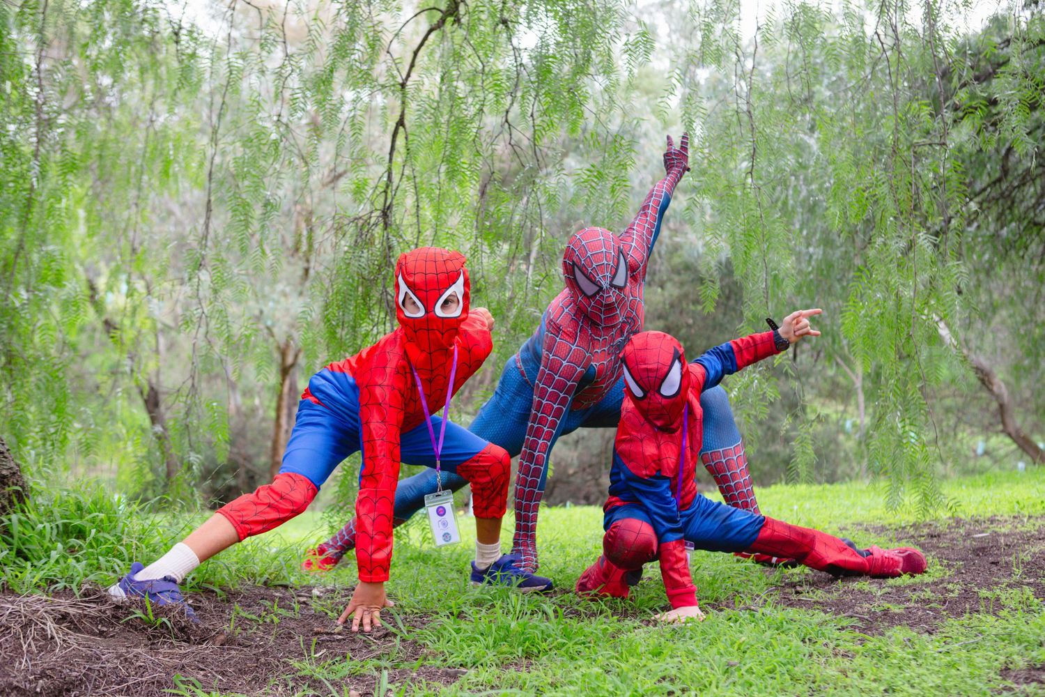 Three children pose in red and blue Spider-Man costumes among green trees outdoors.