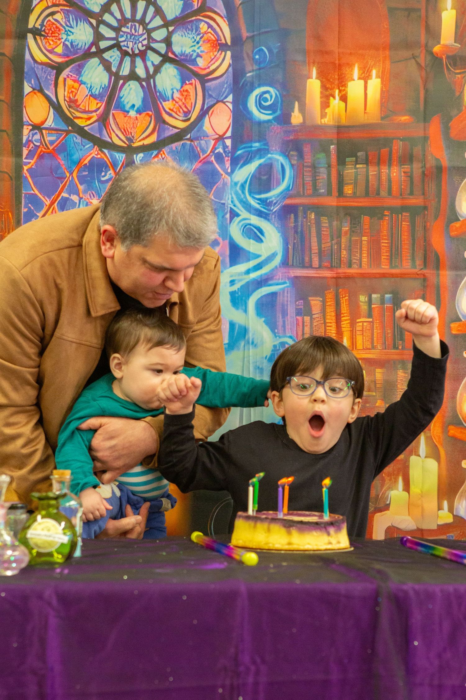 A child with glasses raises fist in excitement before birthday cake while man holds baby, set against colorful fantasy backdrop.
