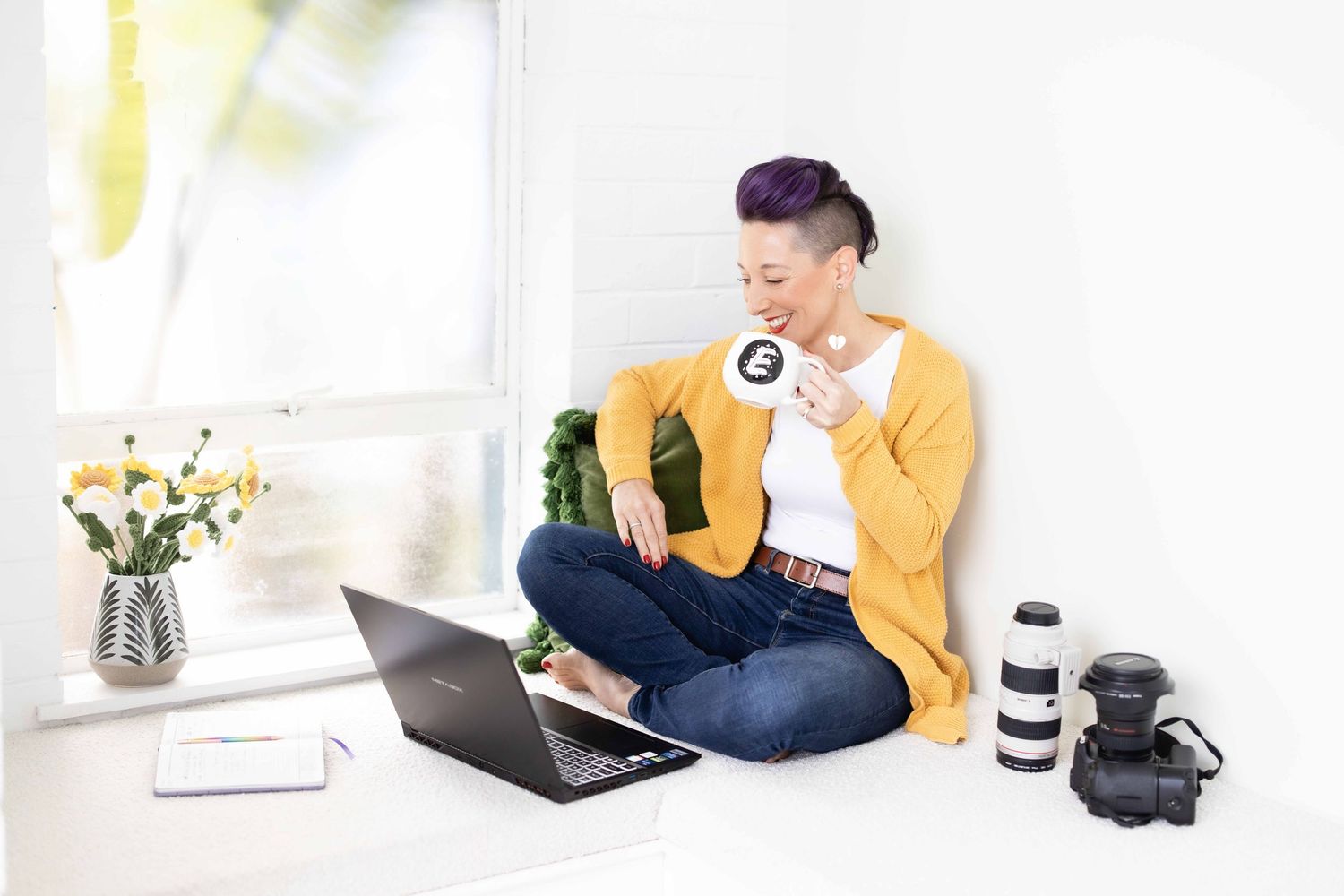 Woman with purple undercut hair in yellow cardigan sitting cross-legged on windowsill with laptop and camera, holding coffee mug.