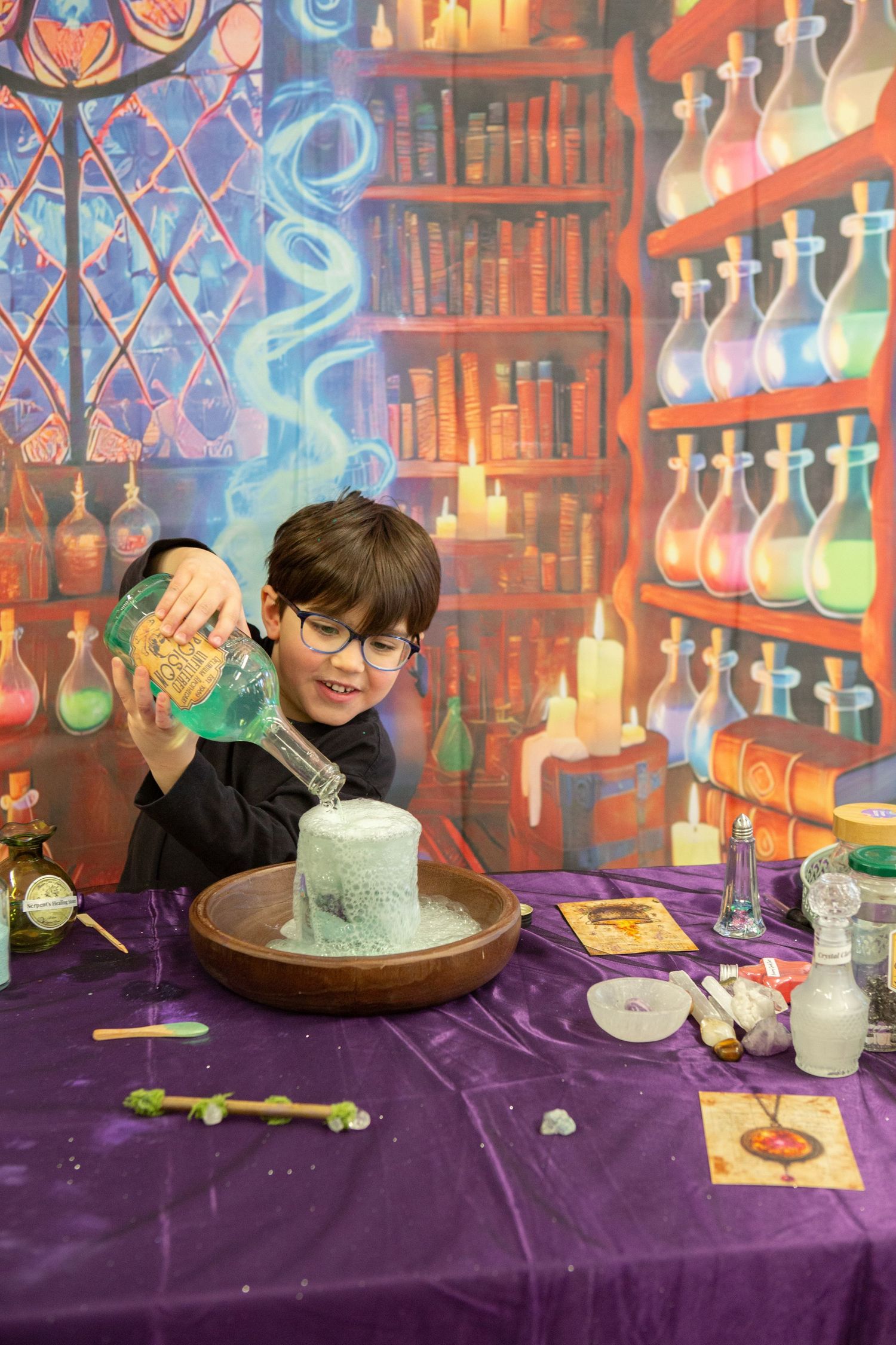 Boy in black wizard robe pouring green liquid into a cauldron at a magical birthday party.