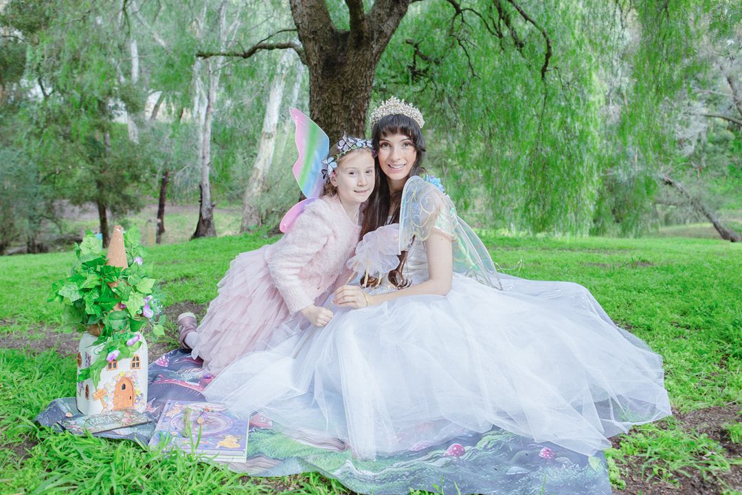Two women in fairy tale costumes with wings and crowns sit together on grass under a willow tree at an outdoor picnic.
