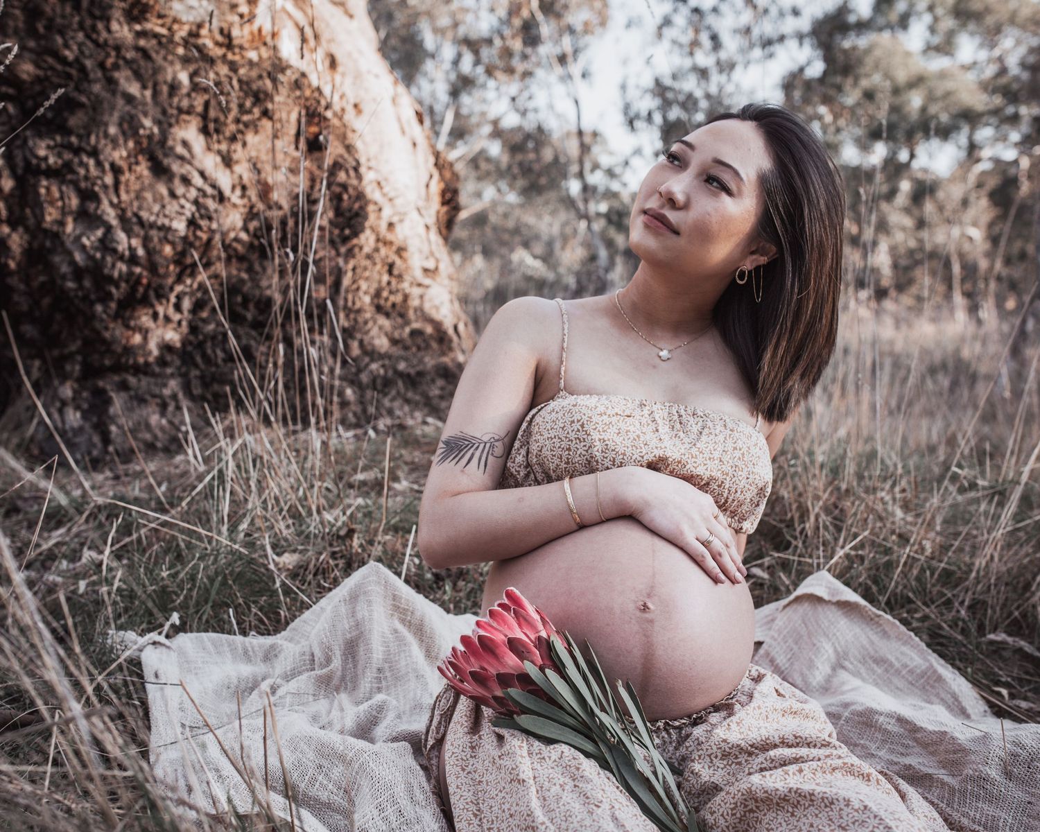 Pregnant woman sitting outdoors in a field holding pink tulips, wearing a floral dress, with natural dry grass background.