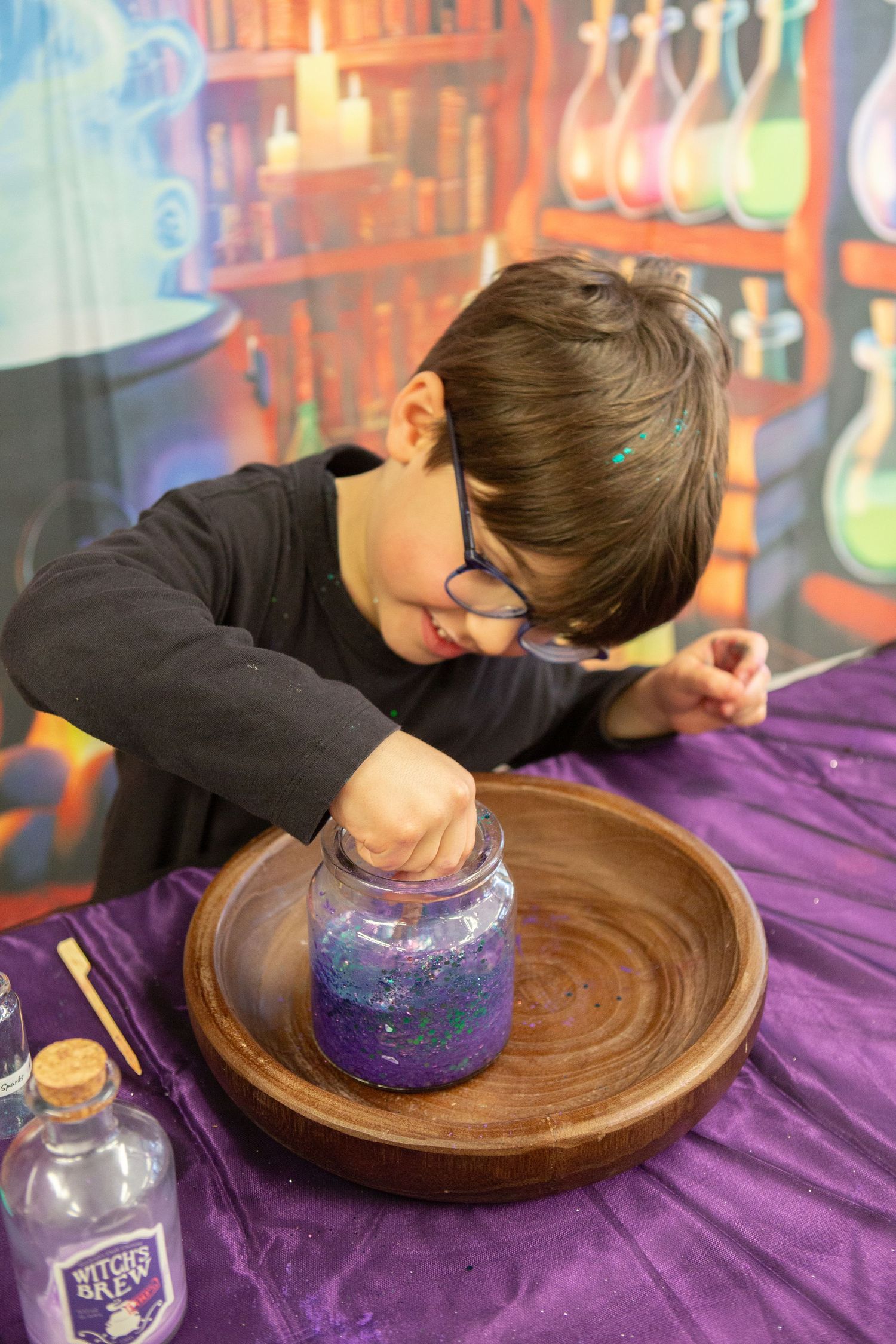 Child with glasses mixing glittery purple potion in jar at wizard-themed craft activity table.
