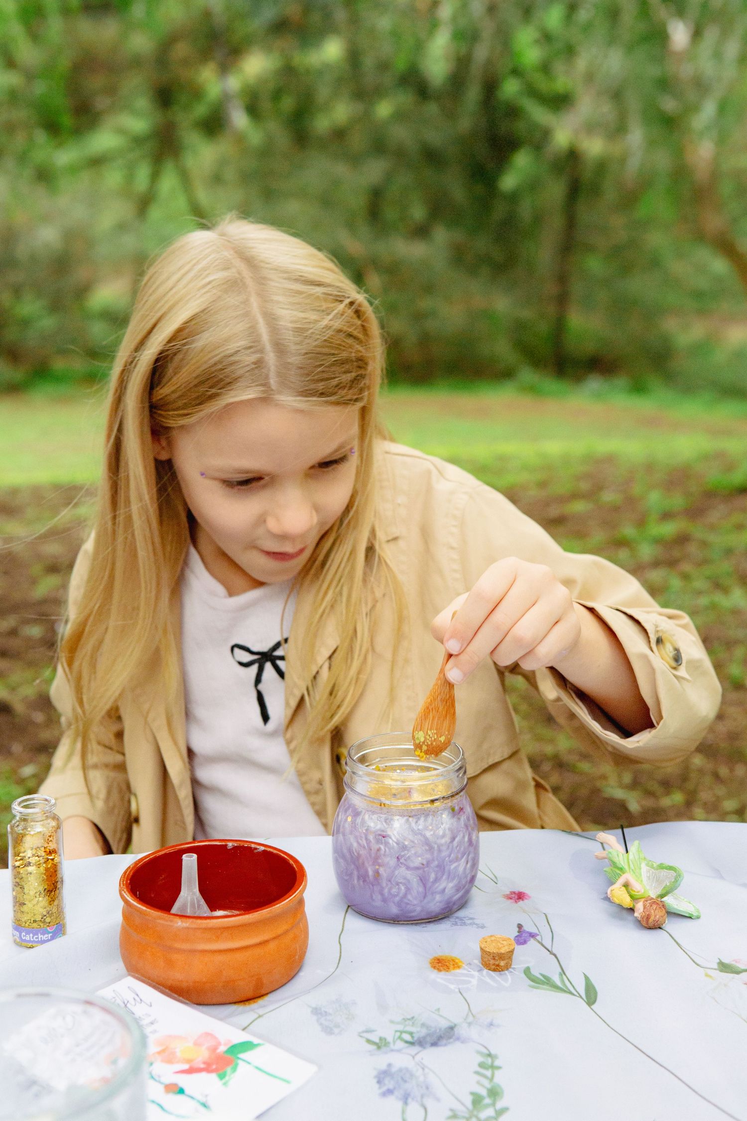 Blonde girl mixing ingredients in a glass jar outdoors, with flowers and spices on a white table.