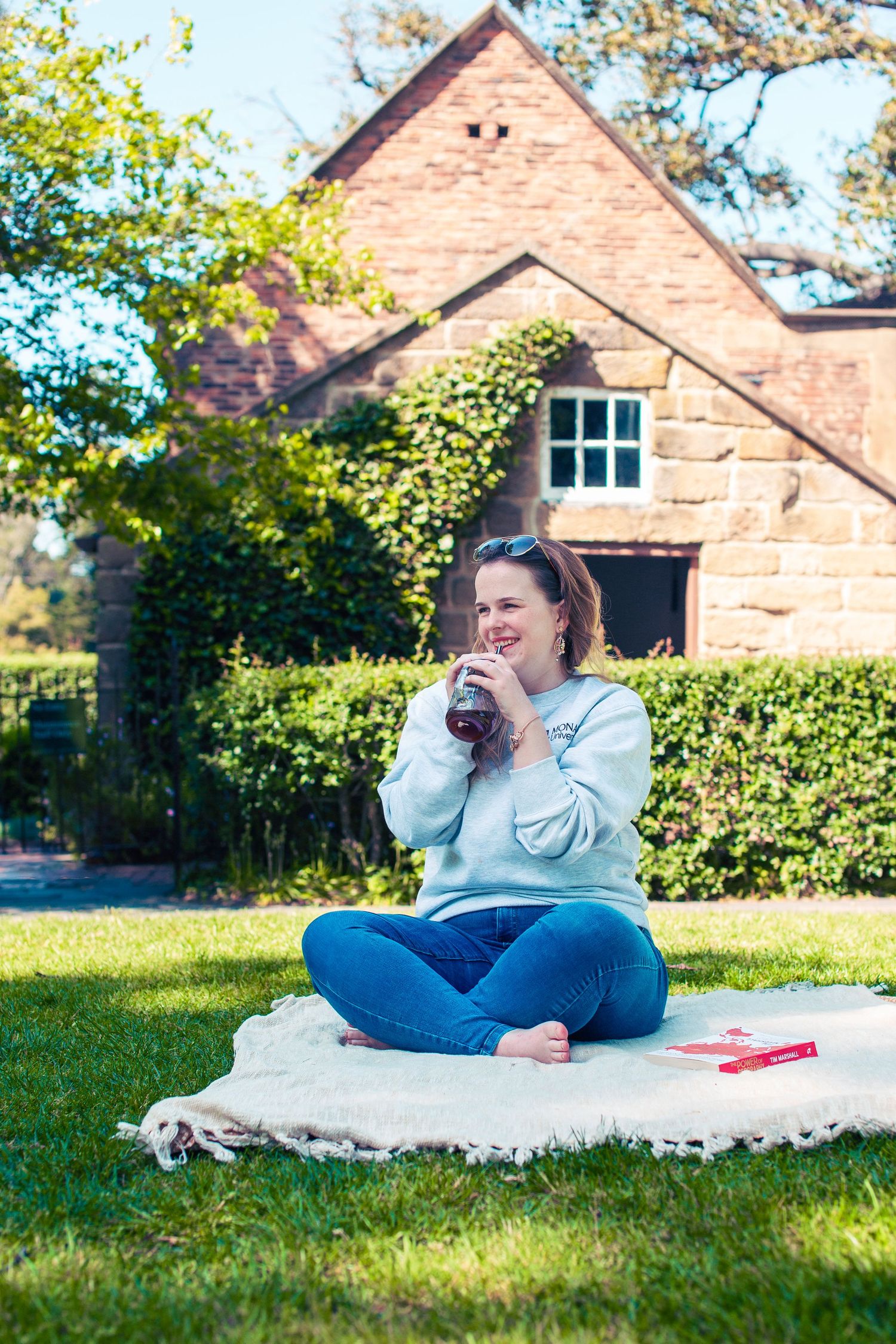Woman sitting cross-legged on blanket in sunny garden, drinking from bottle, wearing grey sweatshirt and jeans near brick house.