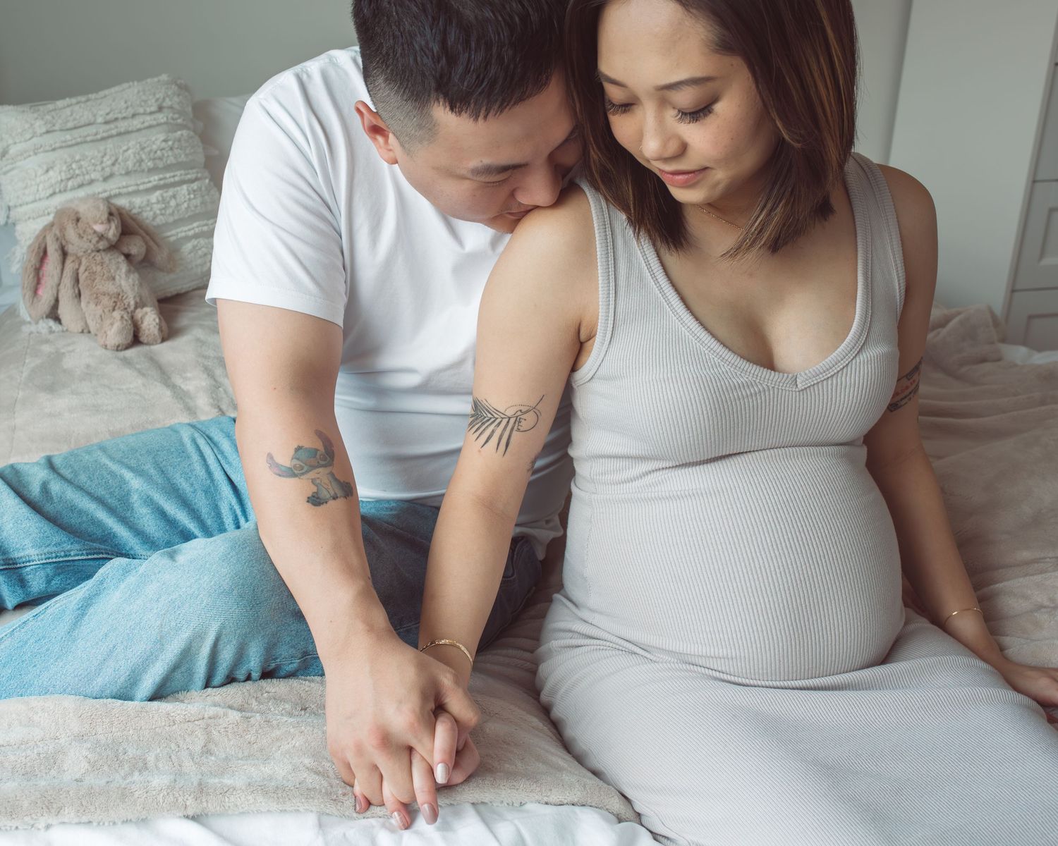 Couple sitting on bed, pregnant woman in gray dress holding hands with tattooed partner in white shirt.