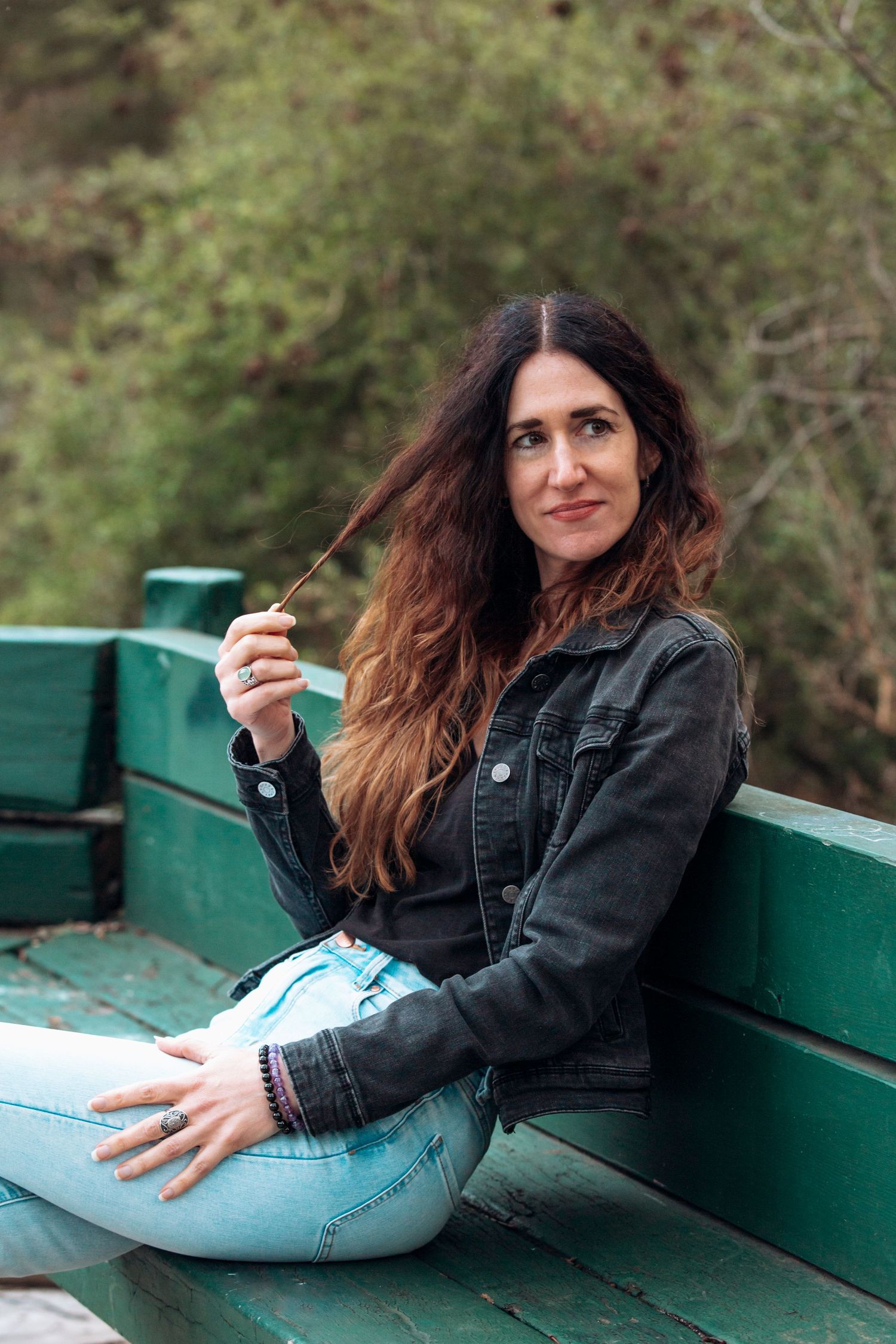 Woman with long wavy hair sitting on a green bench outdoors, wearing a black denim jacket and light blue jeans.