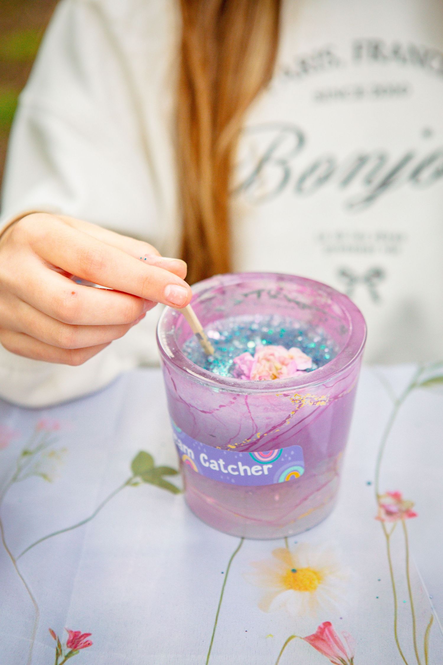 Girl stirring glitter and flowers in a purple Dream Catcher resin craft jar on a floral tablecloth.