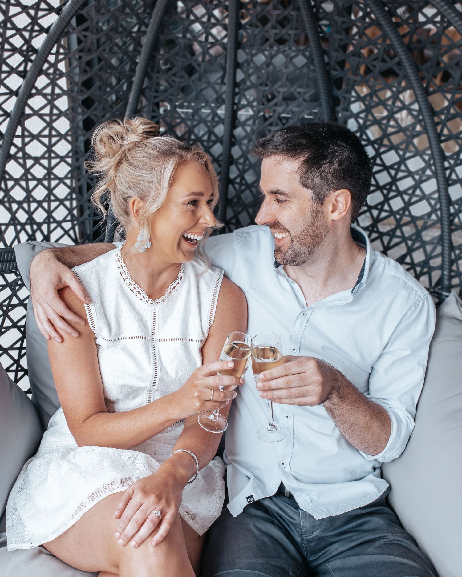 Happy couple toasting with champagne glasses while sitting in a wicker egg chair, smiling at each other.