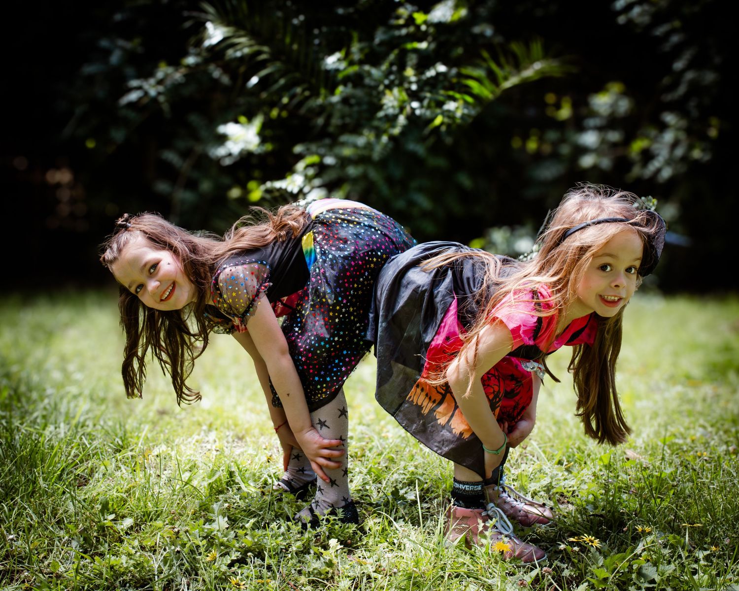 Two young girls in colorful outfits playfully pose back-to-back on a bright green grassy lawn.