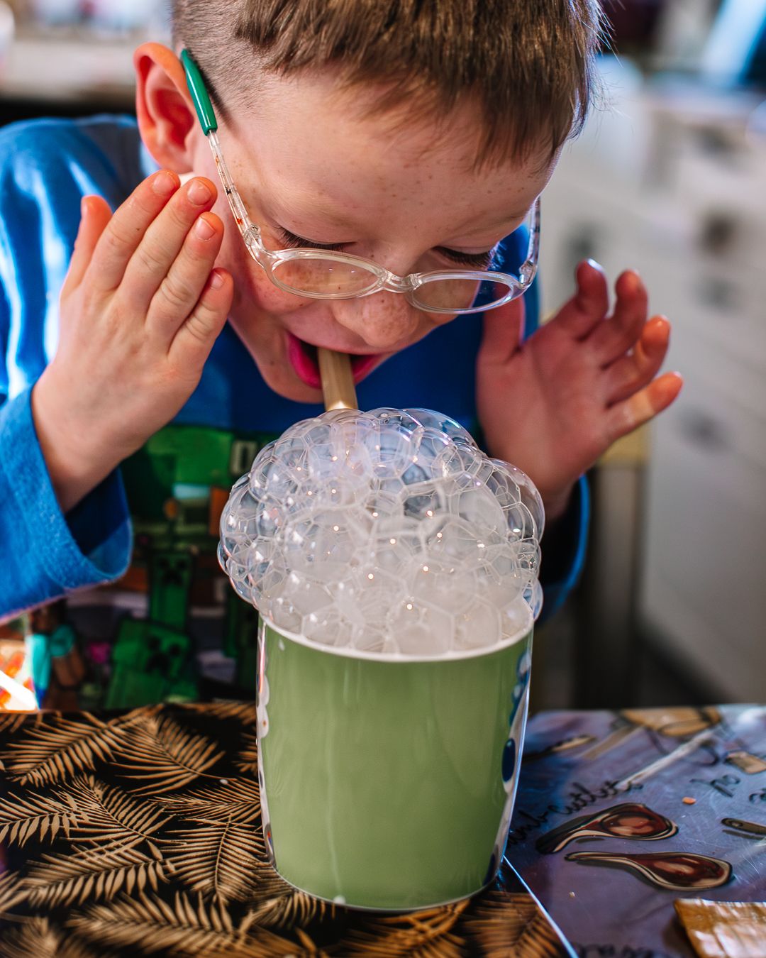 Young boy with glasses excitedly blowing bubbles through a straw in a green cup.