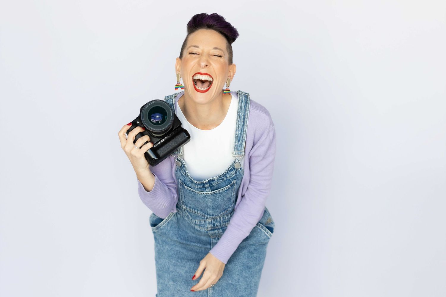 A cheerful woman with short purple hair laughs while holding a DSLR camera against a white background.