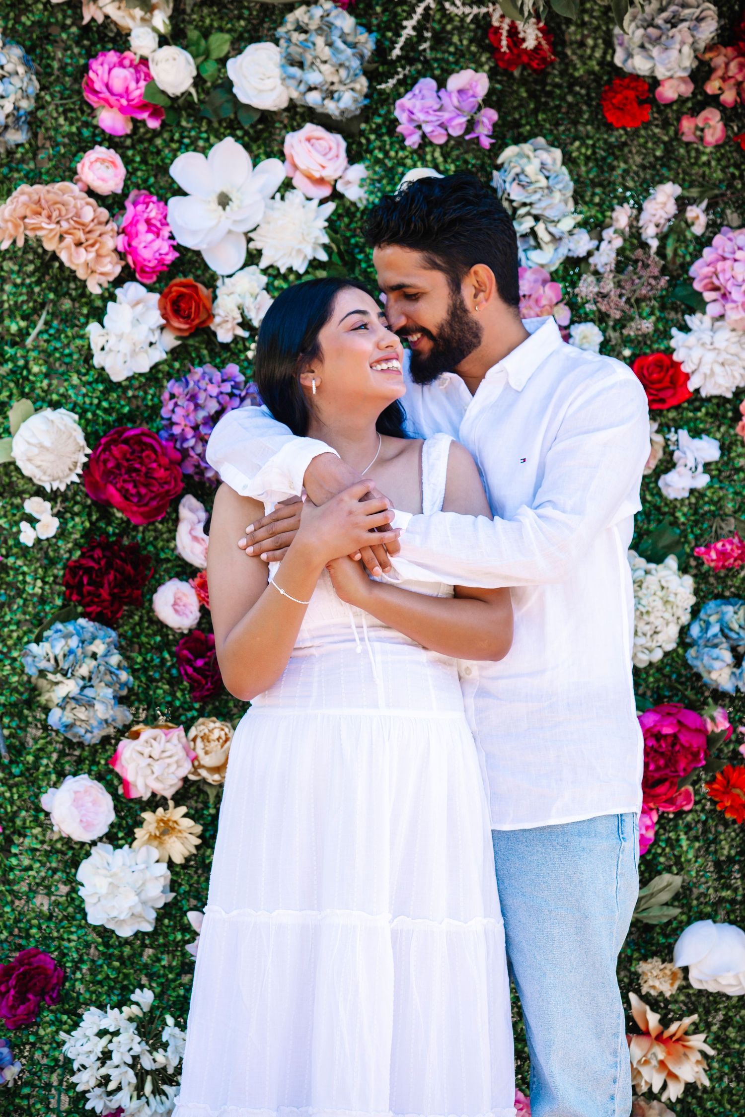 Smiling couple embraces in front of a vibrant colorful flower wall backdrop.