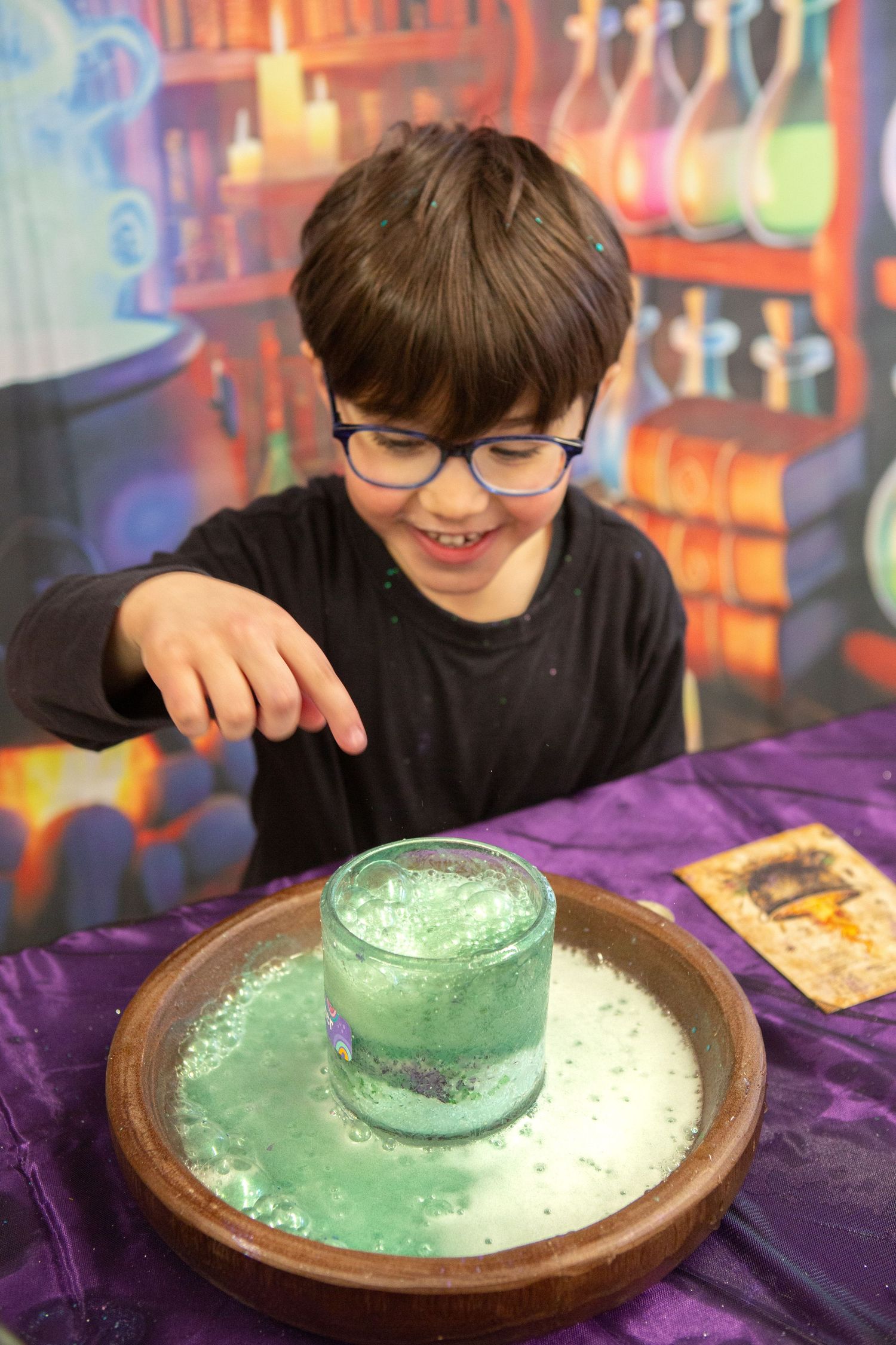 A smiling boy with glasses points at a bubbling green science experiment in a jar on a purple table.