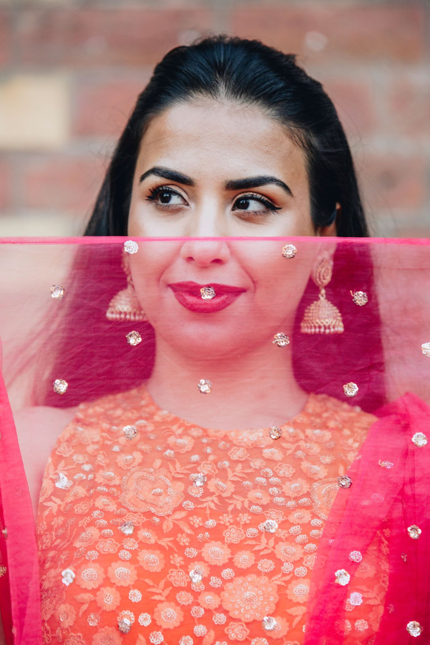A woman in an orange embroidered lehenga with pink dupatta and jhumka earrings poses elegantly.