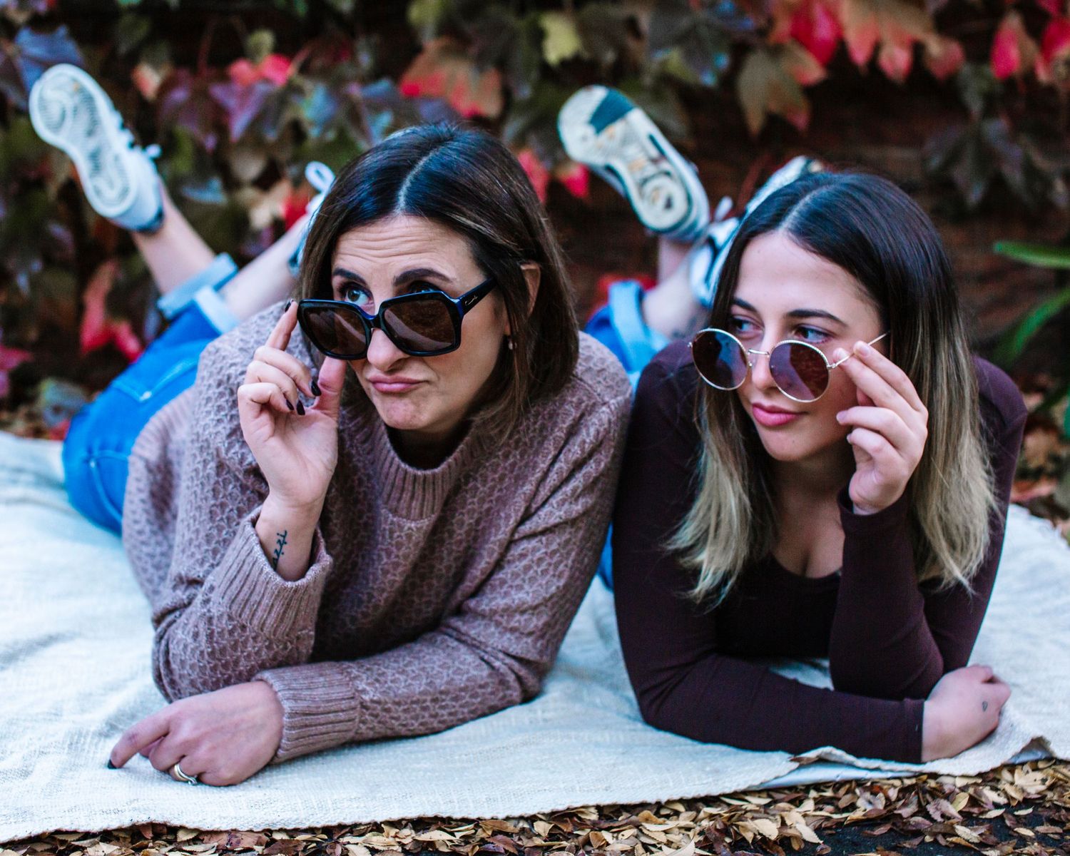 Two young women lying on a blanket outdoors, wearing stylish sunglasses and posing playfully among colorful autumn leaves.