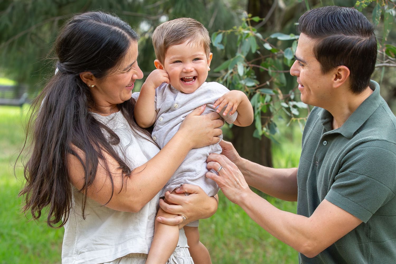 Two parents laugh and play with their happy baby boy in a green outdoor setting.