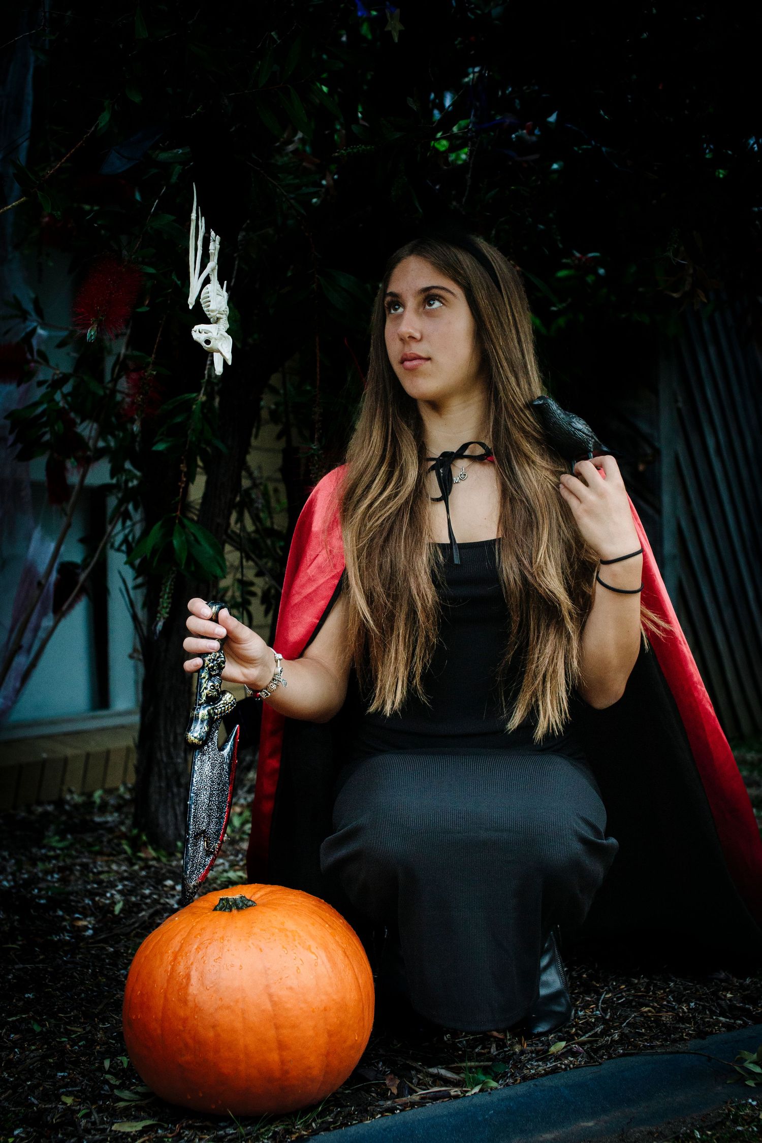 Teen girl in black dress and red cape Halloween costume poses with pumpkin and crow prop outdoors.