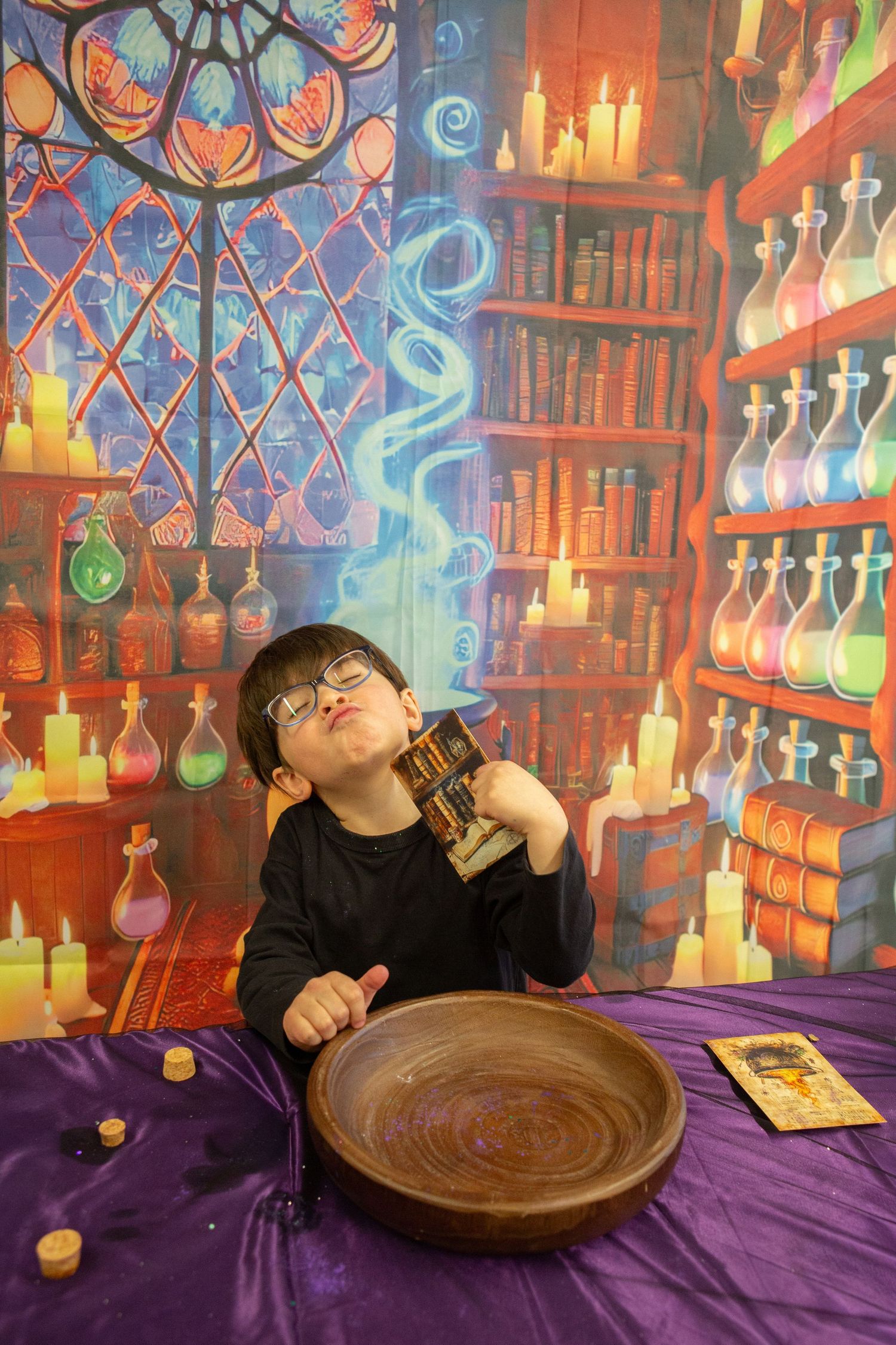 Child in wizard costume holds potion bottle in magical alchemist laboratory backdrop with colorful potions and candles.