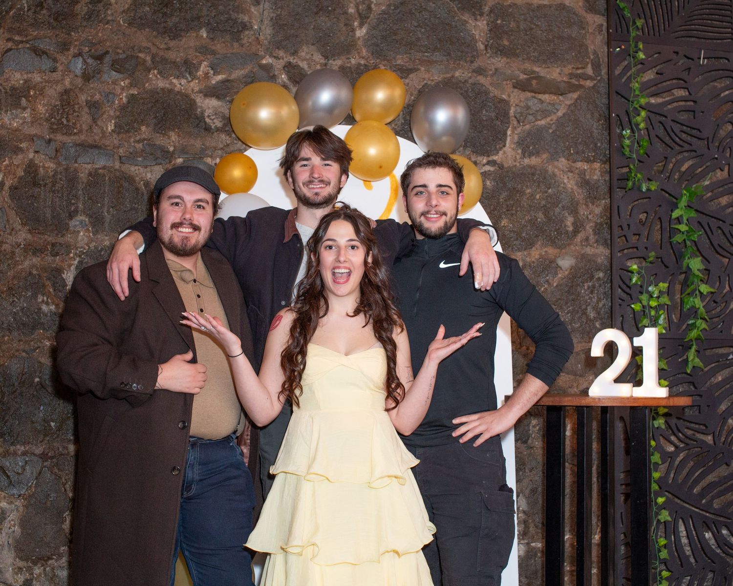 Four young adults posing together at a 21st birthday party with gold and silver balloons and stone wall backdrop.