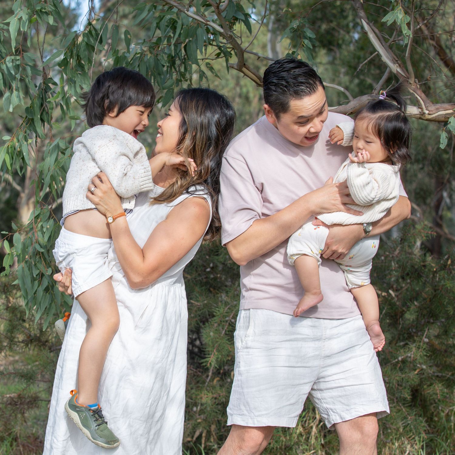 Asian family of four with two young children smiling together outdoors among eucalyptus trees.