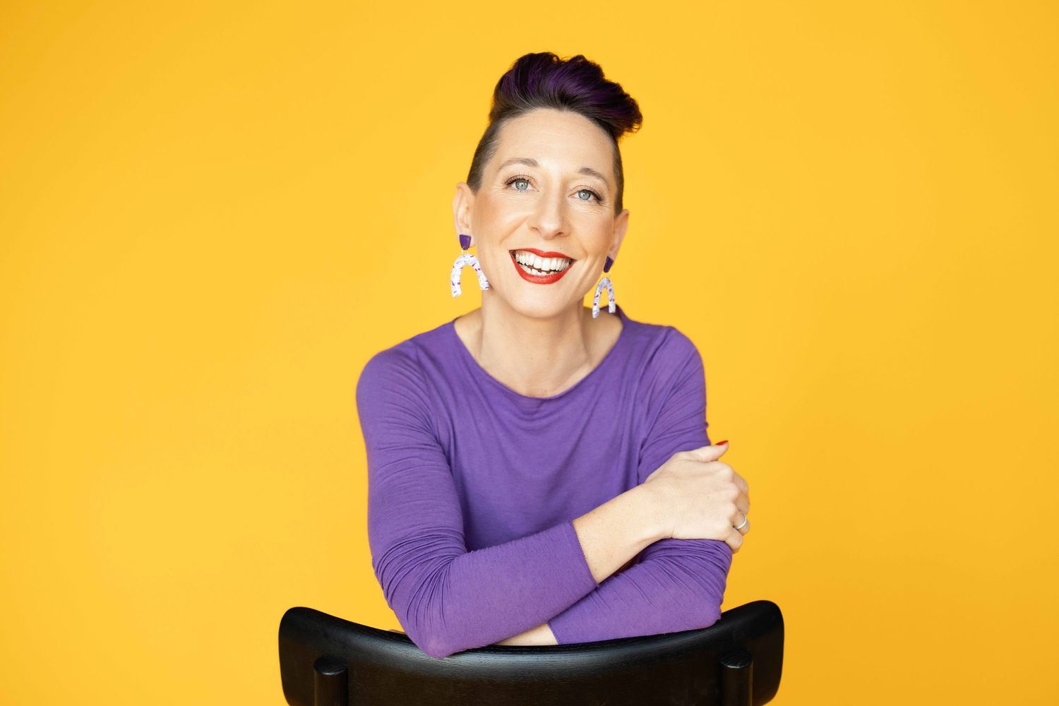 Smiling woman in purple top with dark updo hair leaning on black chair with arms crossed against bright yellow background.