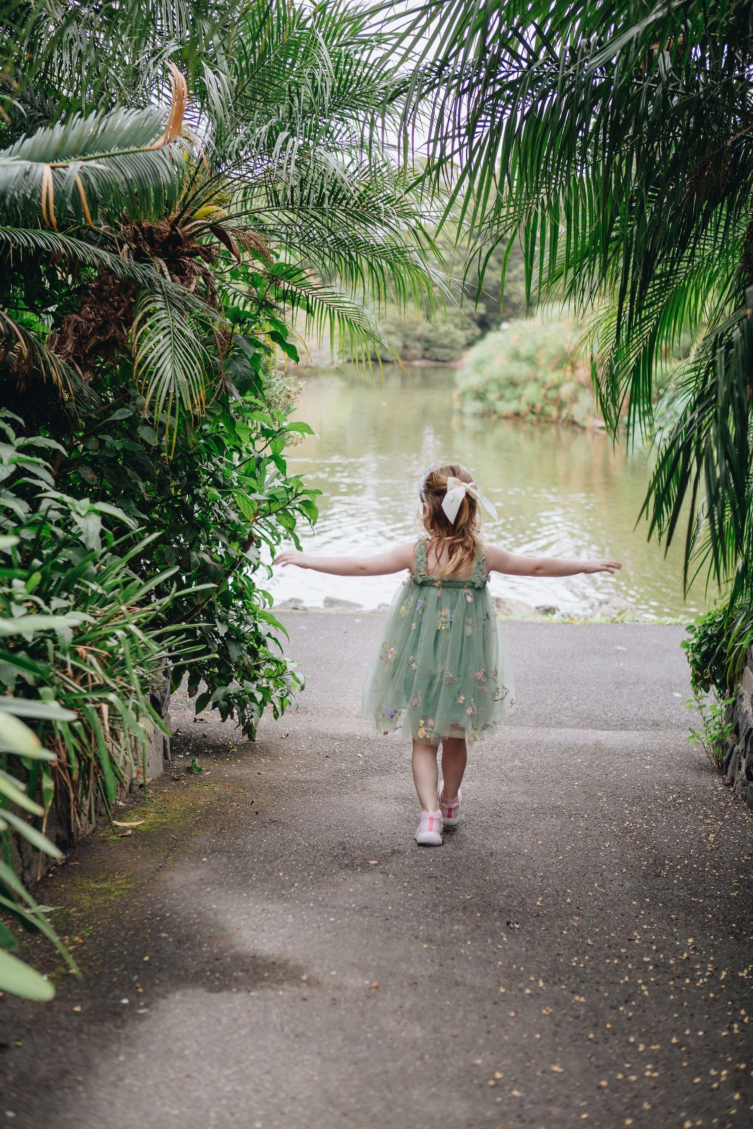 A young girl in a green tulle dress walks with arms outstretched toward a serene pond surrounded by tropical plants.