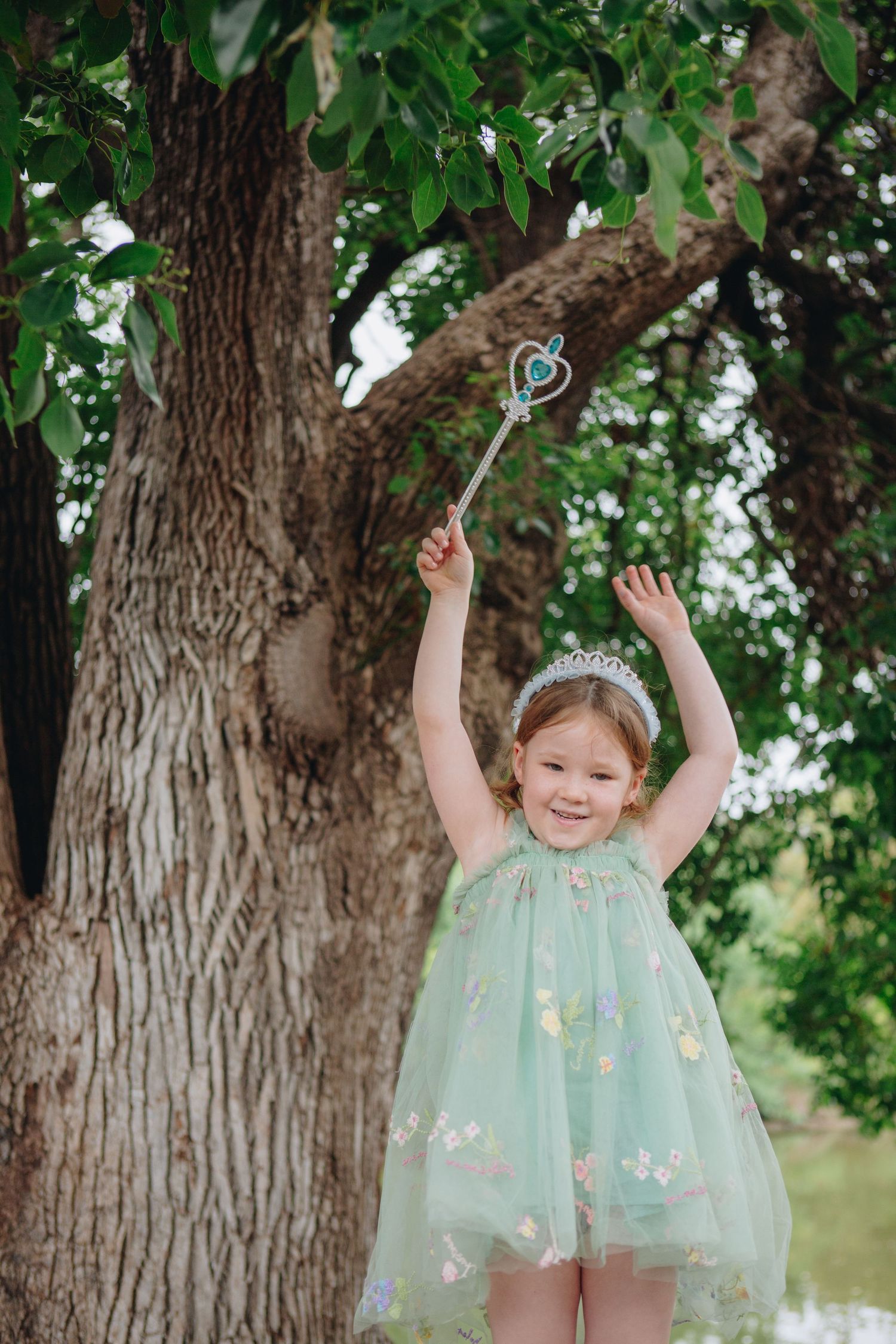Young girl in mint green princess dress holds magic wand aloft, wearing tiara, smiling joyfully beside large tree outdoors.