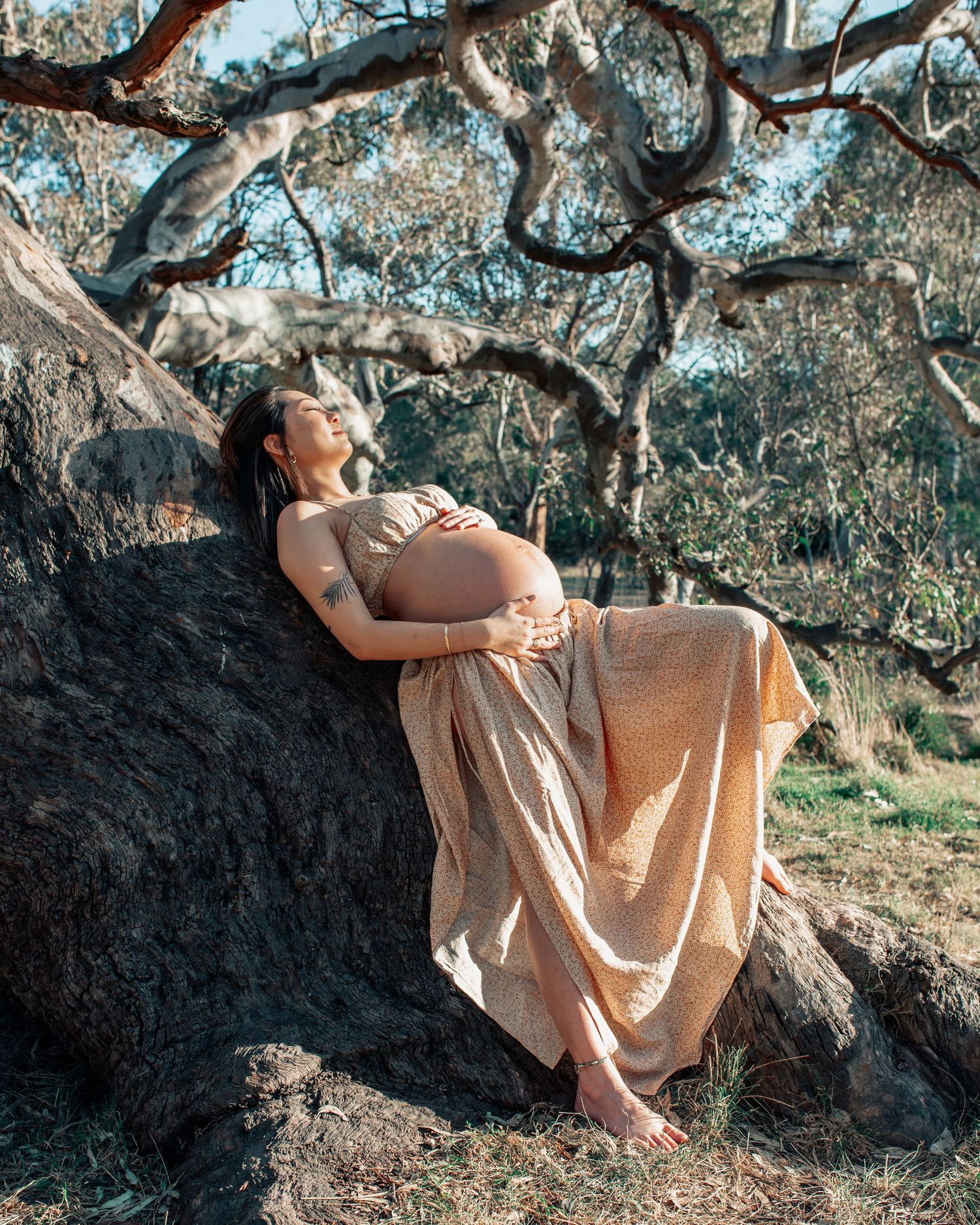 Pregnant woman in beige dress leaning against rocks under gnarled trees, cradling her baby bump outdoors.