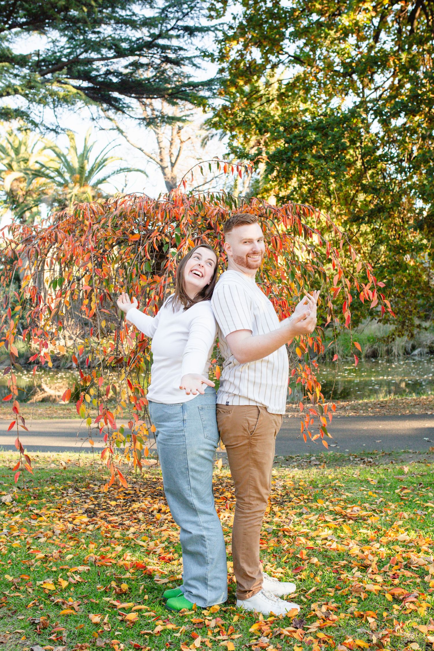 Couple laughing and posing back-to-back under colorful autumn trees in a park with red and orange foliage.