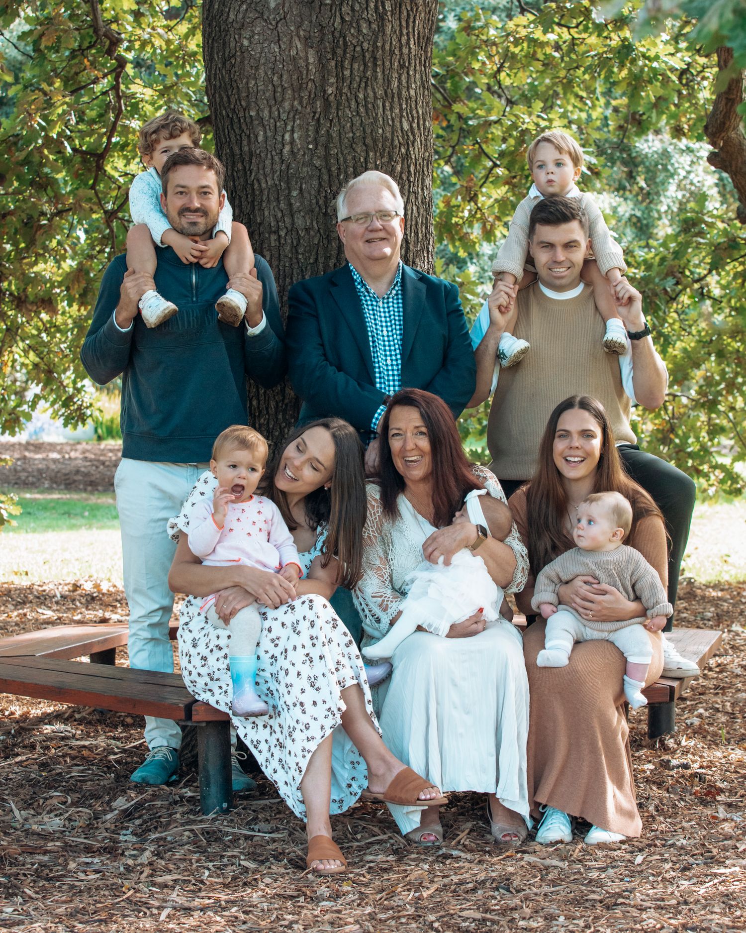 Large multigenerational family portrait with babies and children posed under a tree in a park.