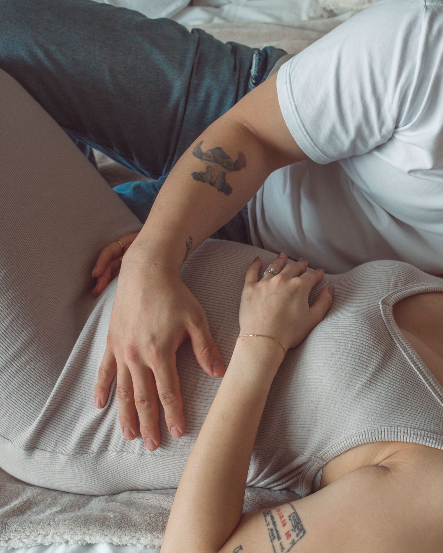 Two people relaxing on a bed, one with tattoos on their arm, wearing casual white and gray clothing.