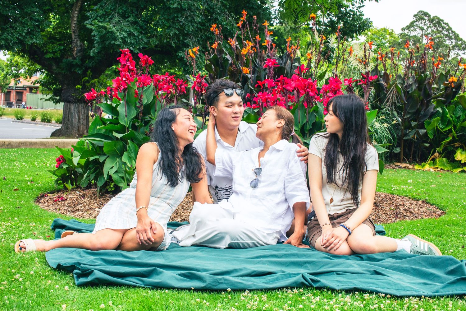 Family of four sitting on a blanket in a lush garden with vibrant red flowers, sharing a joyful moment together.
