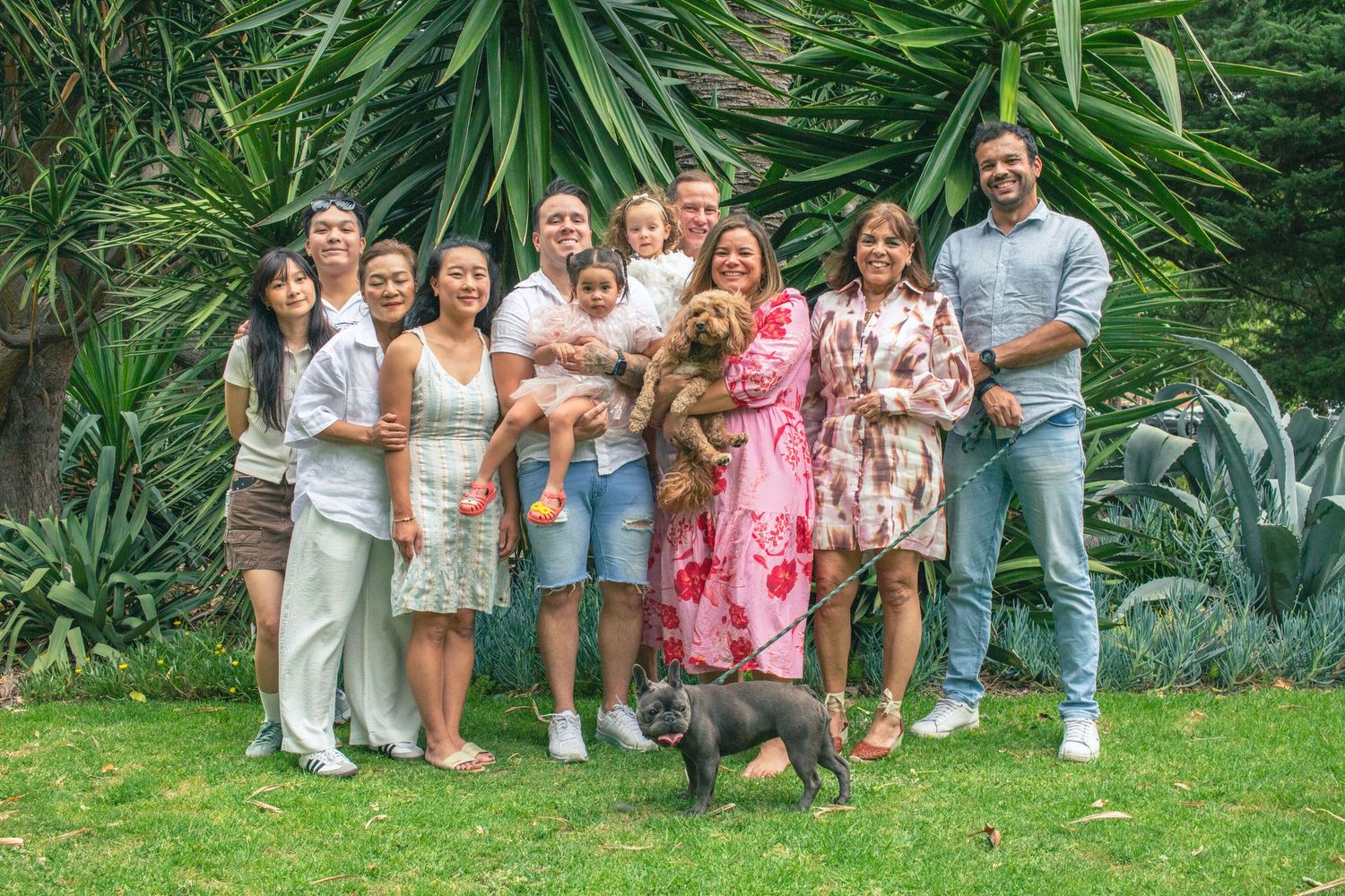 Large family group smiling outdoors in lush garden with two dogs, including a French Bulldog, on a sunny day.