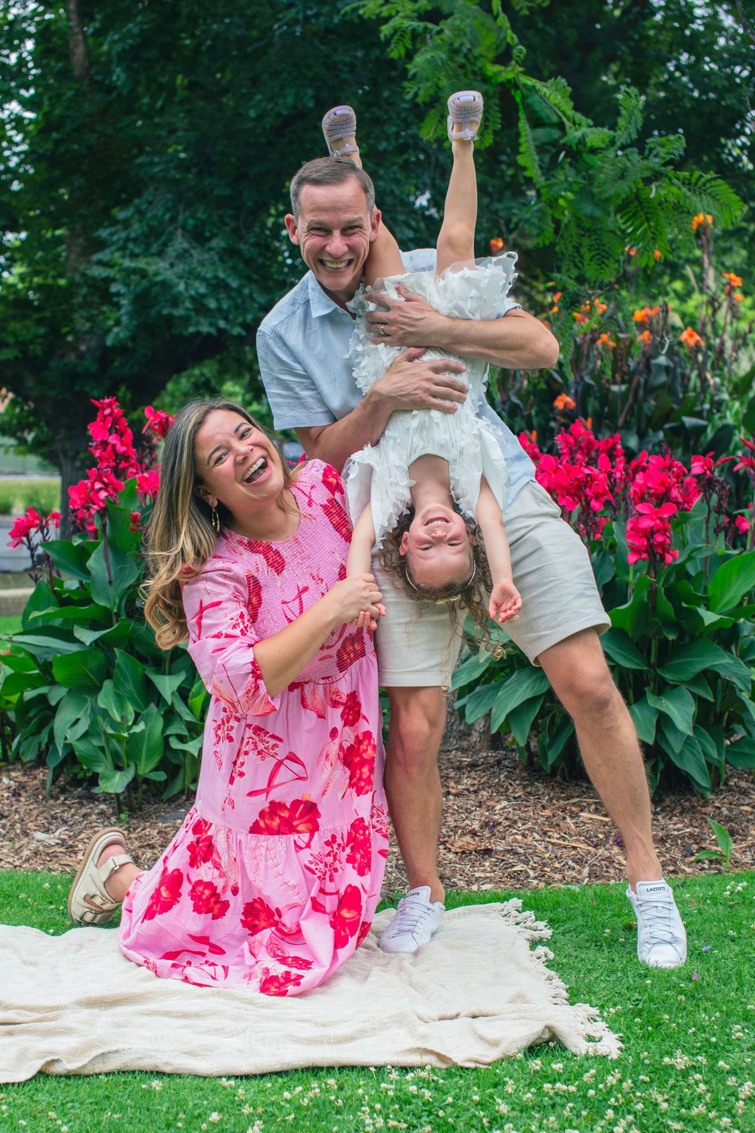 A joyful family laughing in a garden, man holding child upside down, woman in pink floral dress, red flowers behind.