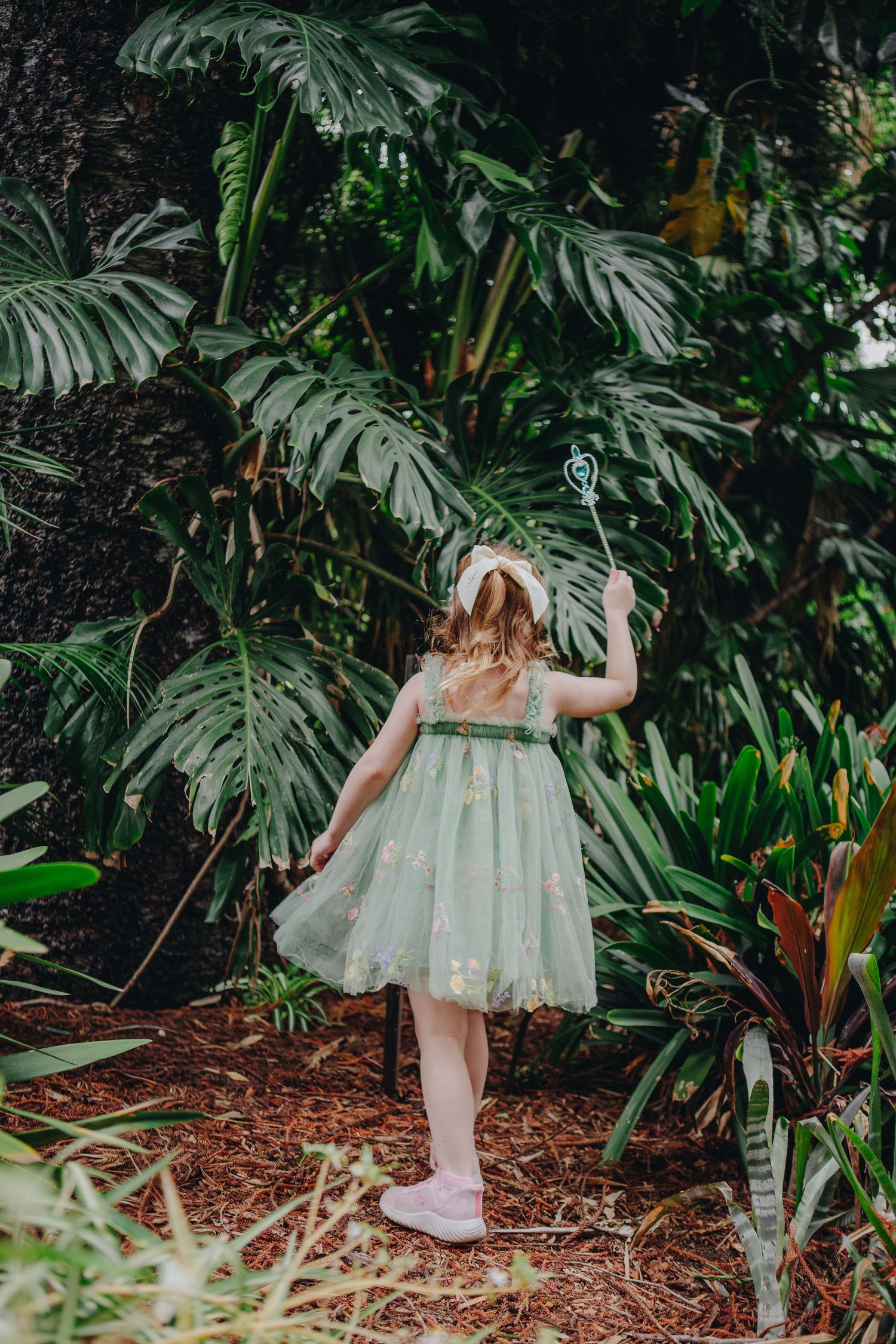 Young girl in green tulle dress holding a wand explores a lush tropical garden with large leafy plants.