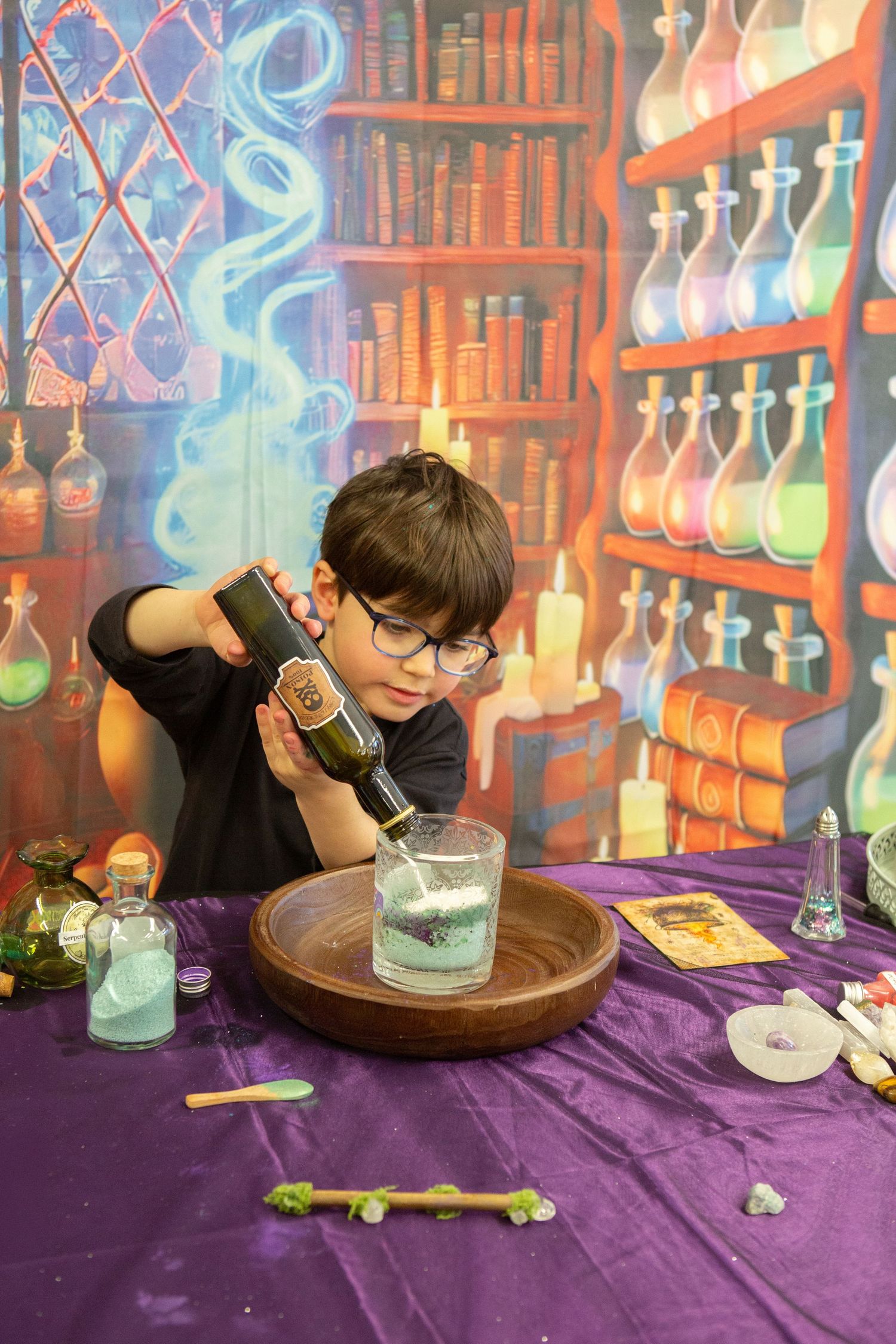 Boy with glasses pouring potion into glass at magic wizard themed table with colorful bookshelf backdrop.