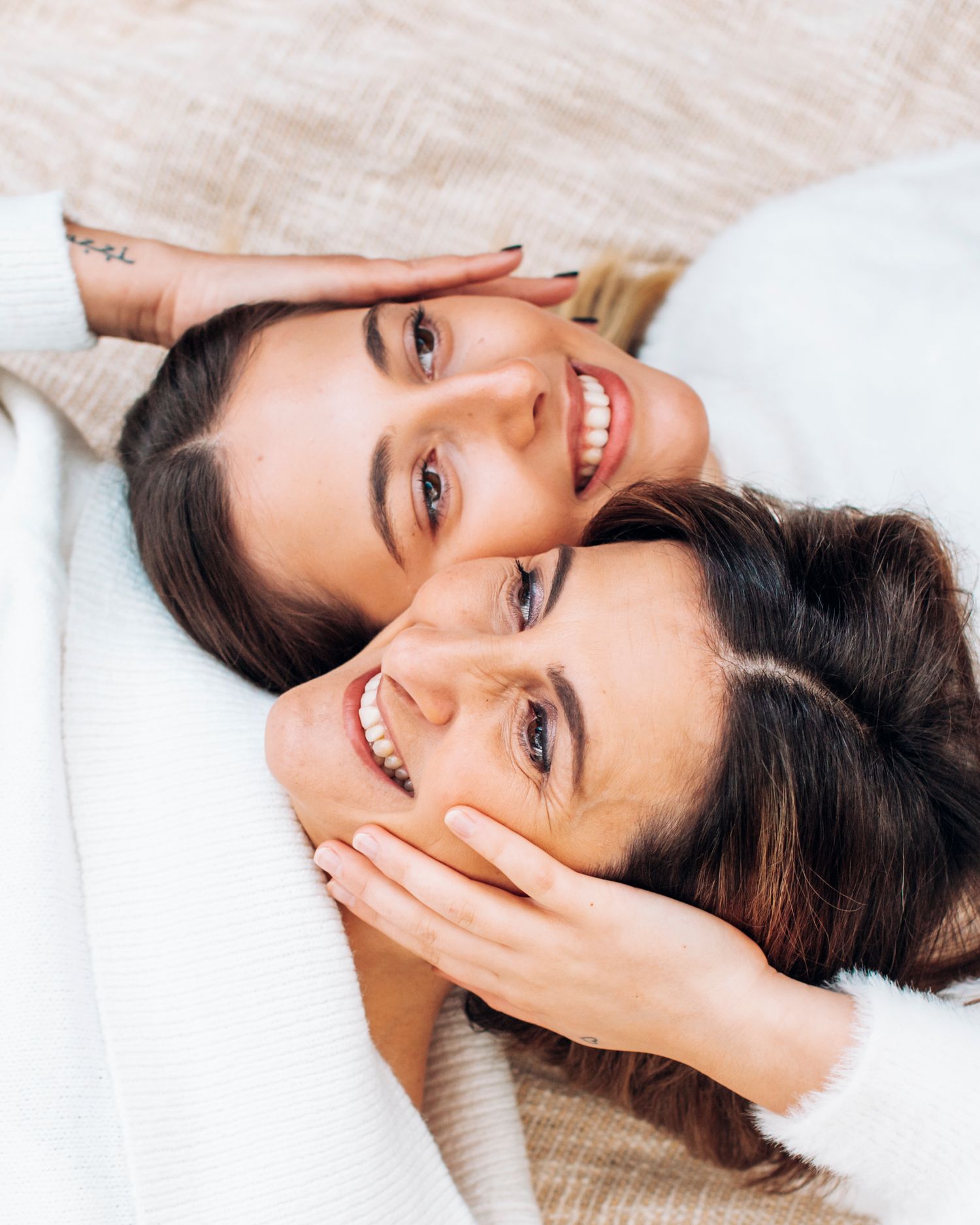 Two smiling brunette women lying down together, cheeks touching, wearing cozy white sweaters in a warm, intimate portrait.