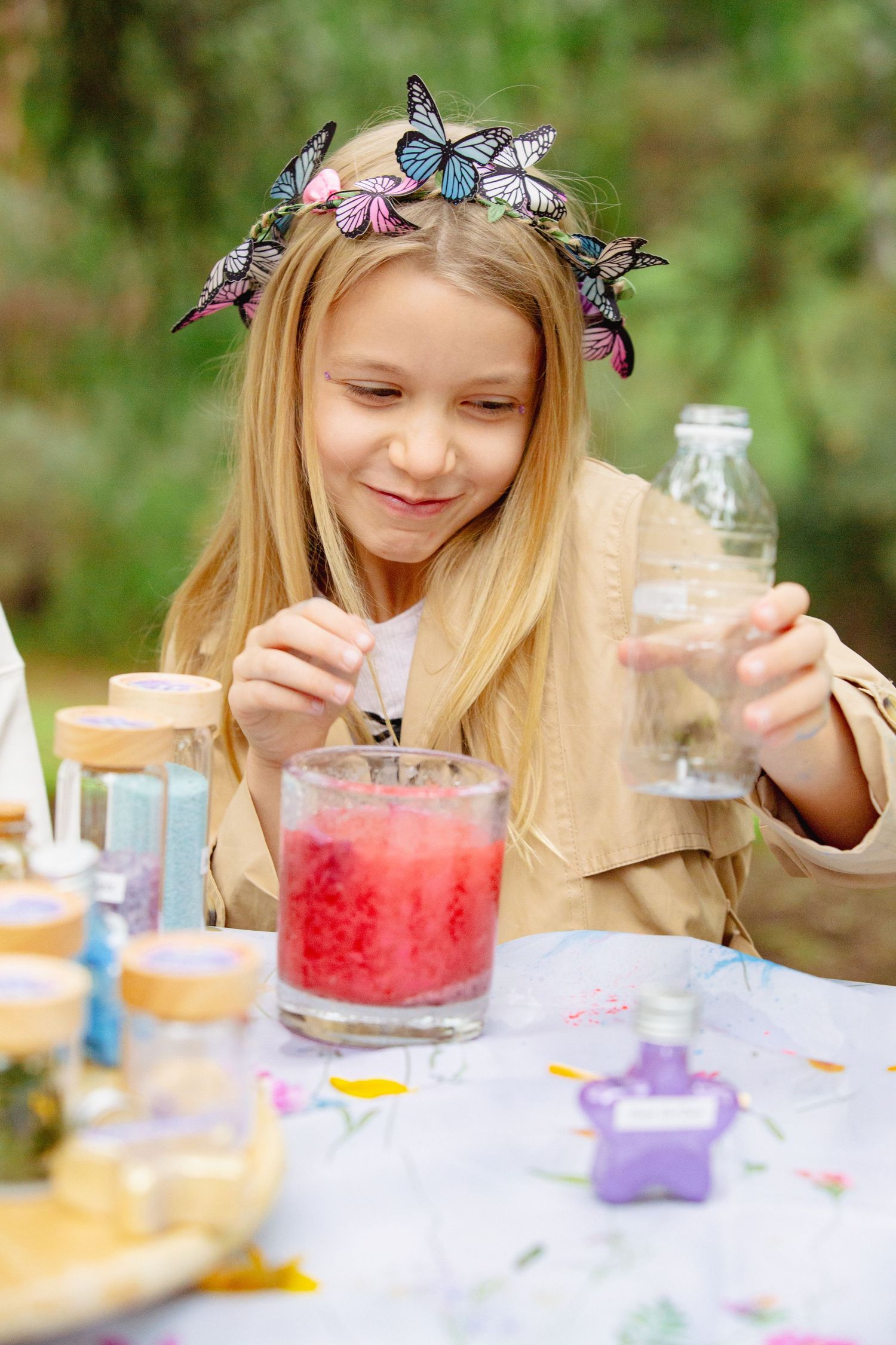 Young girl with butterfly crown mixing colorful potions at outdoor table with small bottles and jars.