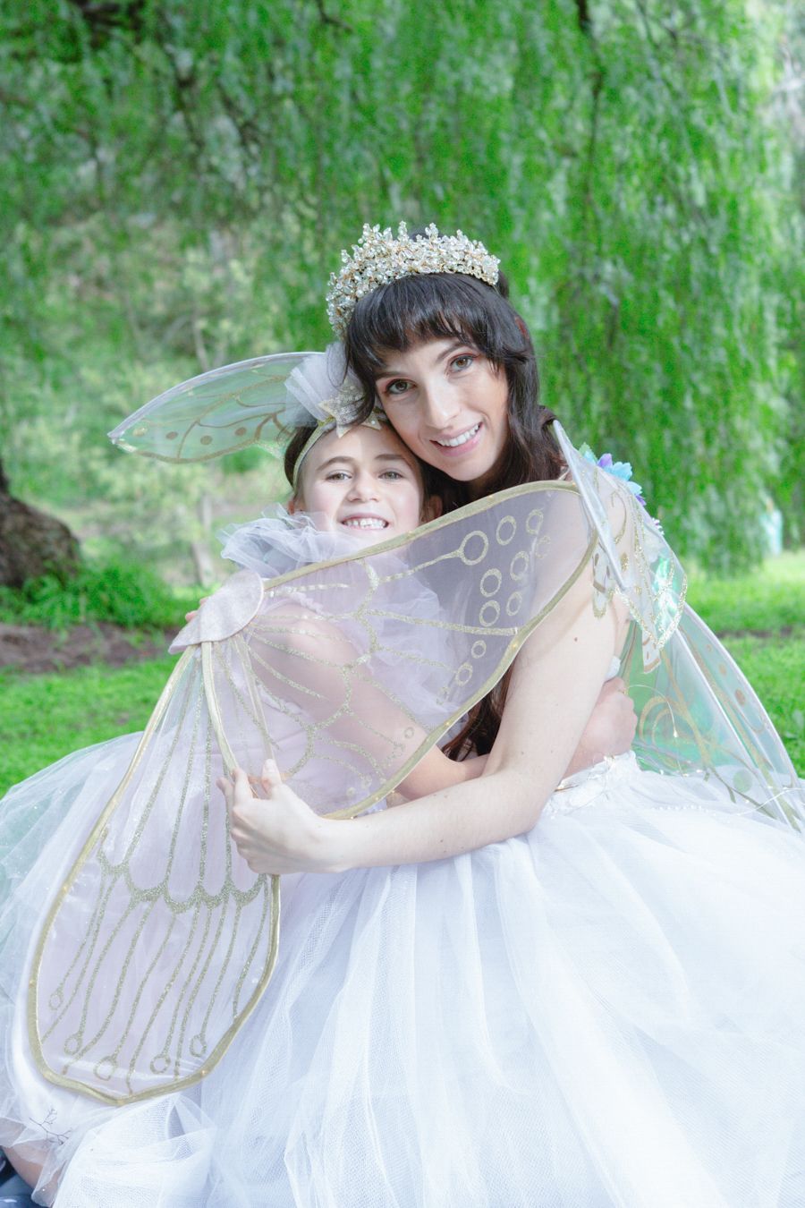Woman and girl in white fairy costumes with wings and tiara hugging outdoors under green willow tree.
