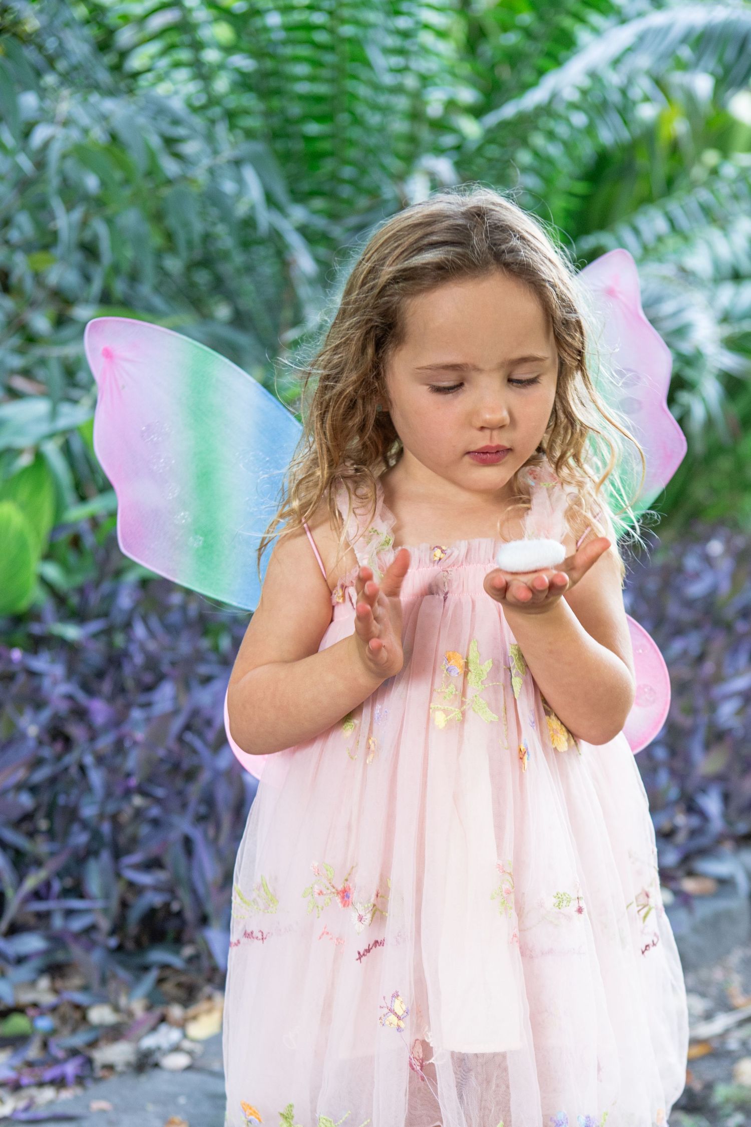 Young girl in pink fairy dress with colorful wings holds a small object in a lush garden setting.