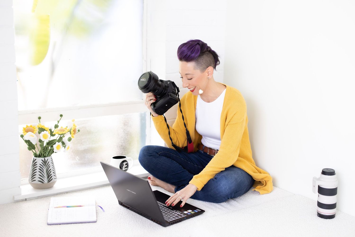 Woman with purple hair sitting cross-legged reviewing photos on a camera and laptop.