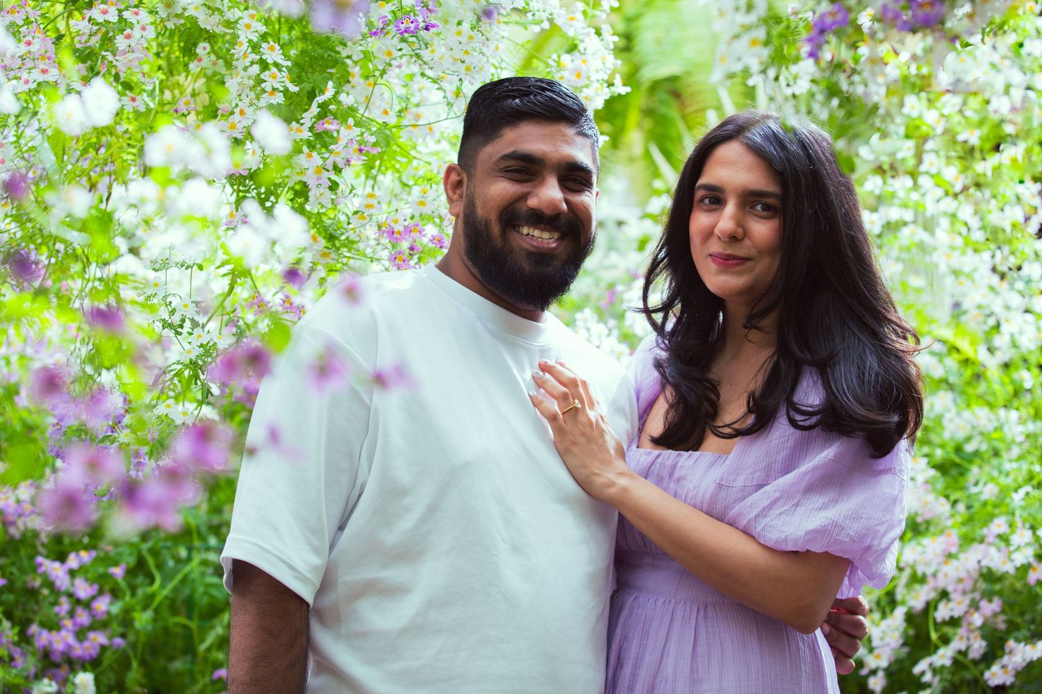 Couple smiling together surrounded by blooming pink and white flowers in a lush garden setting.