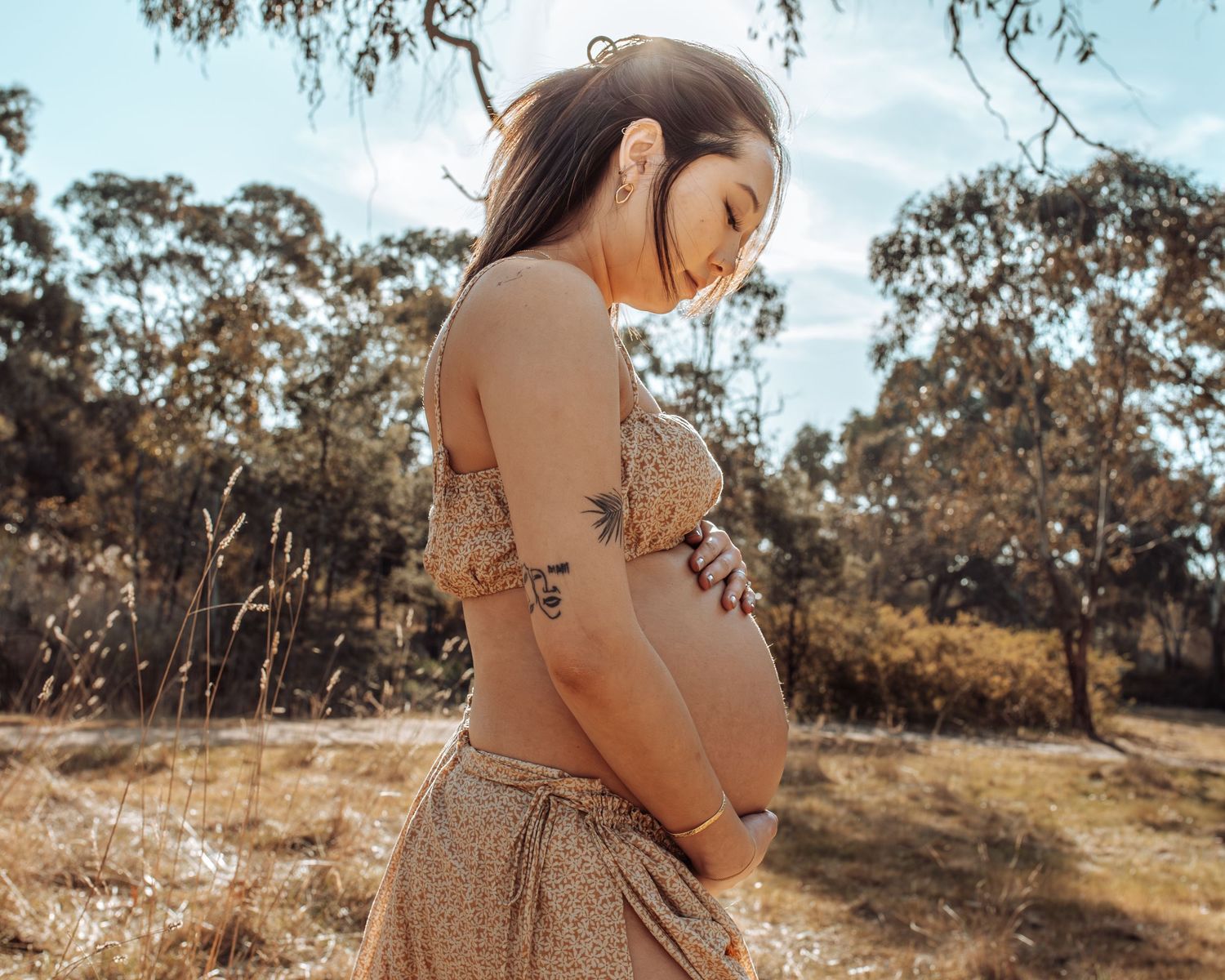Pregnant woman in floral skirt cradling her baby bump outdoors in a sunlit natural setting.