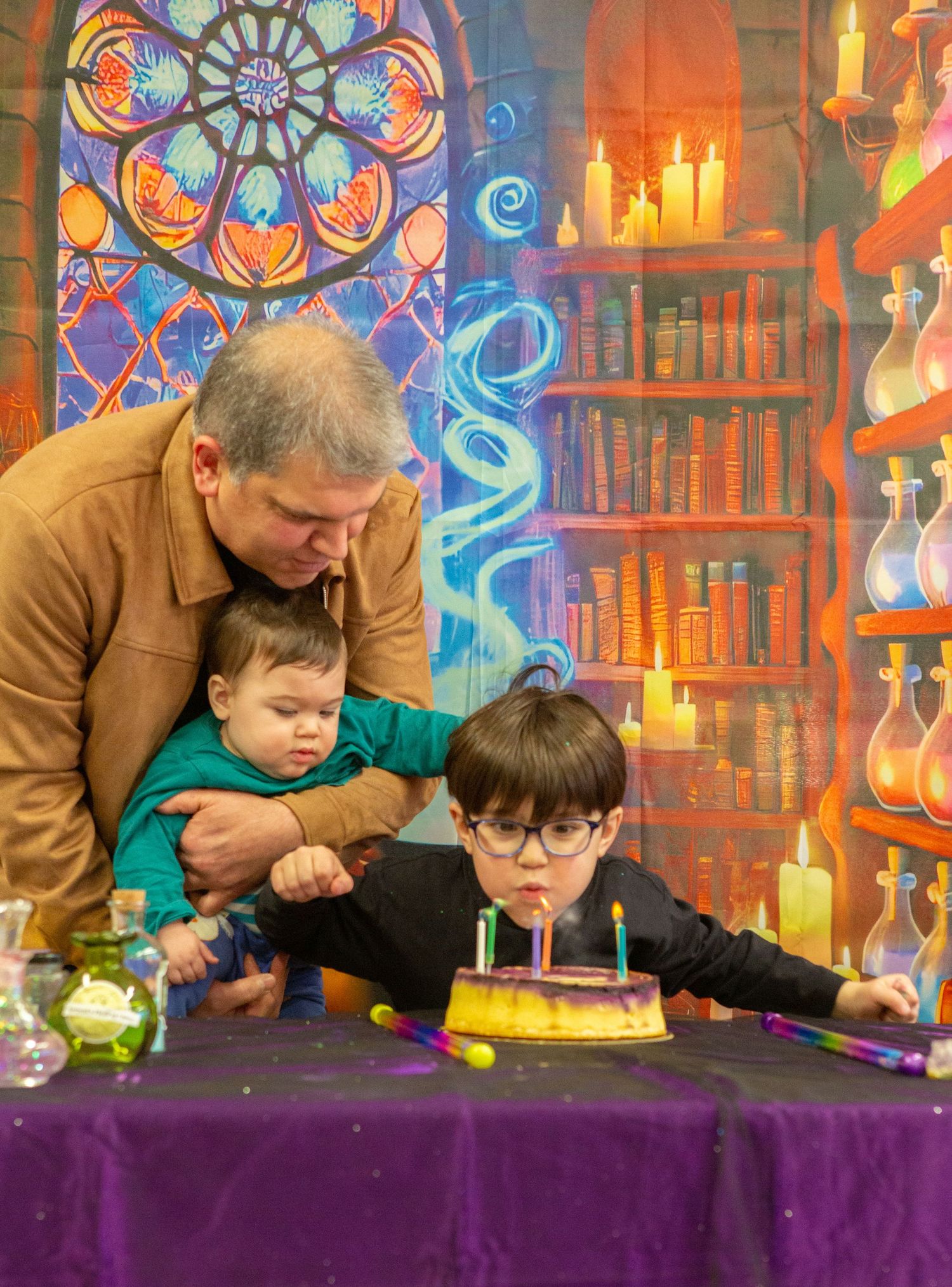 Father holds baby while boy blows birthday candles in magical wizard laboratory themed party setting.