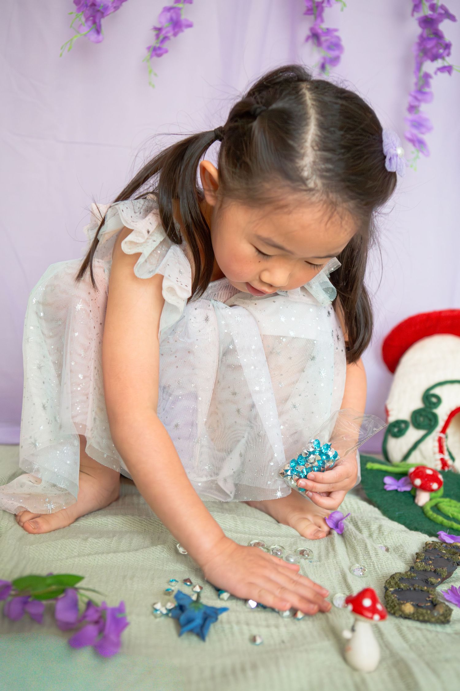 Young Asian girl in white tulle dress playing with fairy garden accessories including mushrooms and flowers.