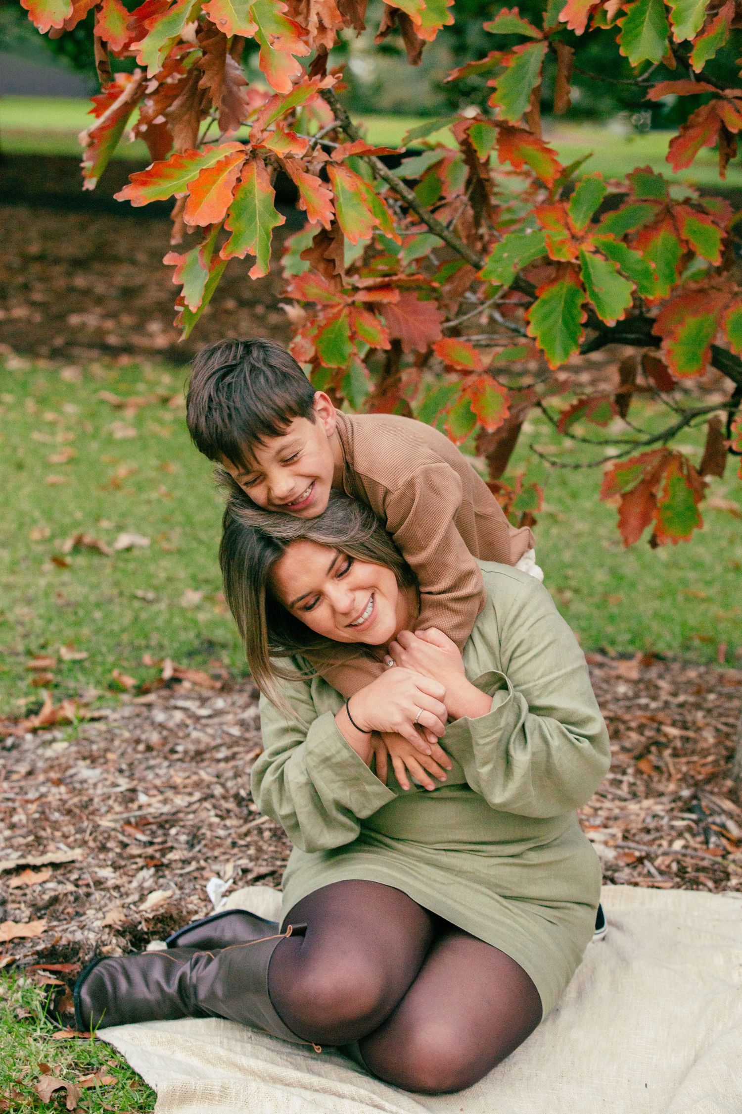 A mother and child share a joyful hug outdoors under colorful autumn leaves in a park.