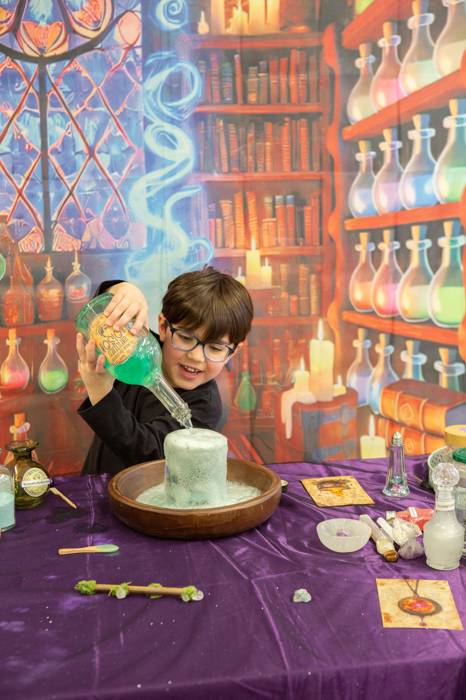 Boy in wizard costume pouring green potion over dry ice cauldron with magical bookshelf backdrop.